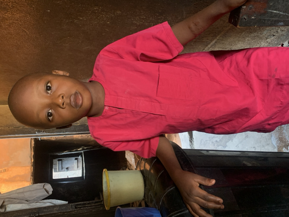 A young Black boy, approximately 4-7 years old, stands centered in what appears to be an indoor residential or communal space in Okene, Nigeria. He has a bald head and a neutral expression, looking directly into the camera with dark eyes and slightly parted lips. He wears a short-sleeved, bright pink tunic with a mandarin collar, a vertical placket, and a left breast pocket. His left hand rests on a dark, curved, possibly upholstered surface, while his right hand rests on a dark, angular, possibly wooden post or door frame.
In the background to the boy's left, a light-colored garment is hanging. Below this, a doorway opens into a darker interior, revealing a smaller, rectangular opening or window further back, through which vague structural elements are discernible. Two plastic buckets, one light yellow with a green rim and another dark blue, are visible on a surface in the lower-left background. The walls in the immediate background are rough-textured and exhibit earthy tones of brown and beige. The scene is illuminated by ambient light, with a brighter light source implied from the distant opening.