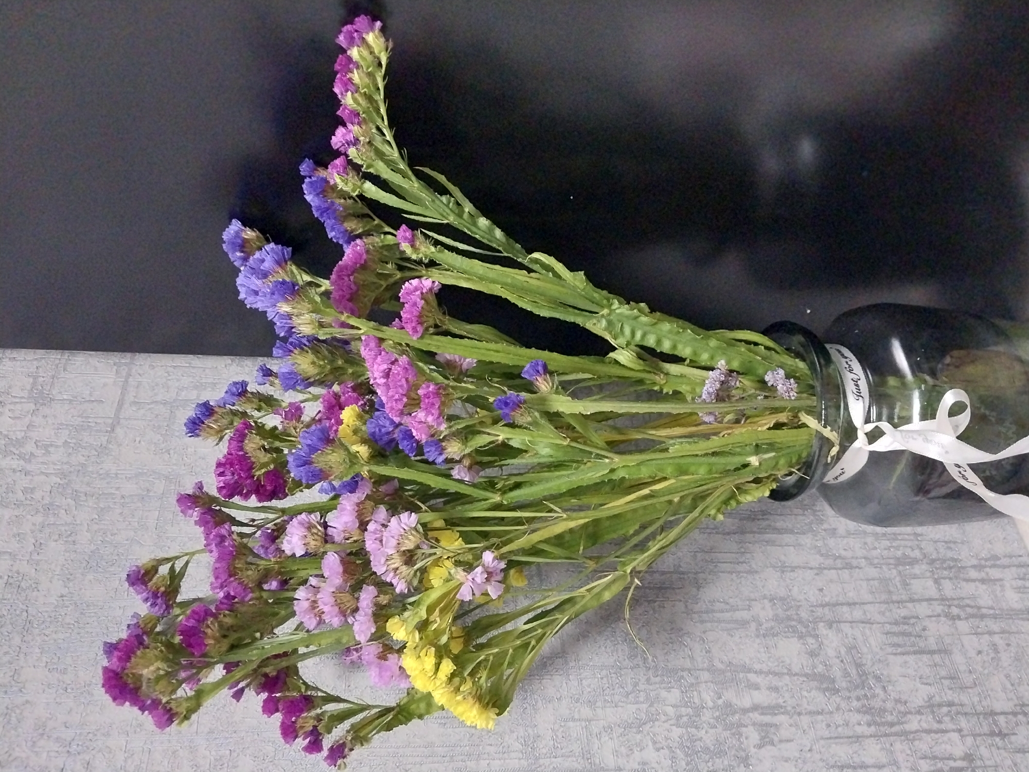 A bouquet of dried flowers, predominantly in shades of purple, pink, and yellow, is arranged in a dark glass vase. The vase is tied with a white ribbon that has the inscription "Just for you" on it. The background consists of a textured gray wall on the left and a dark, out-of-focus area on the right. The lighting is moderate, casting soft shadows. The scene is devoid of any human presence or activity. The image appears to be a still life. There are no discernible features to indicate a specific location within the city of Dnipro, Ukraine.