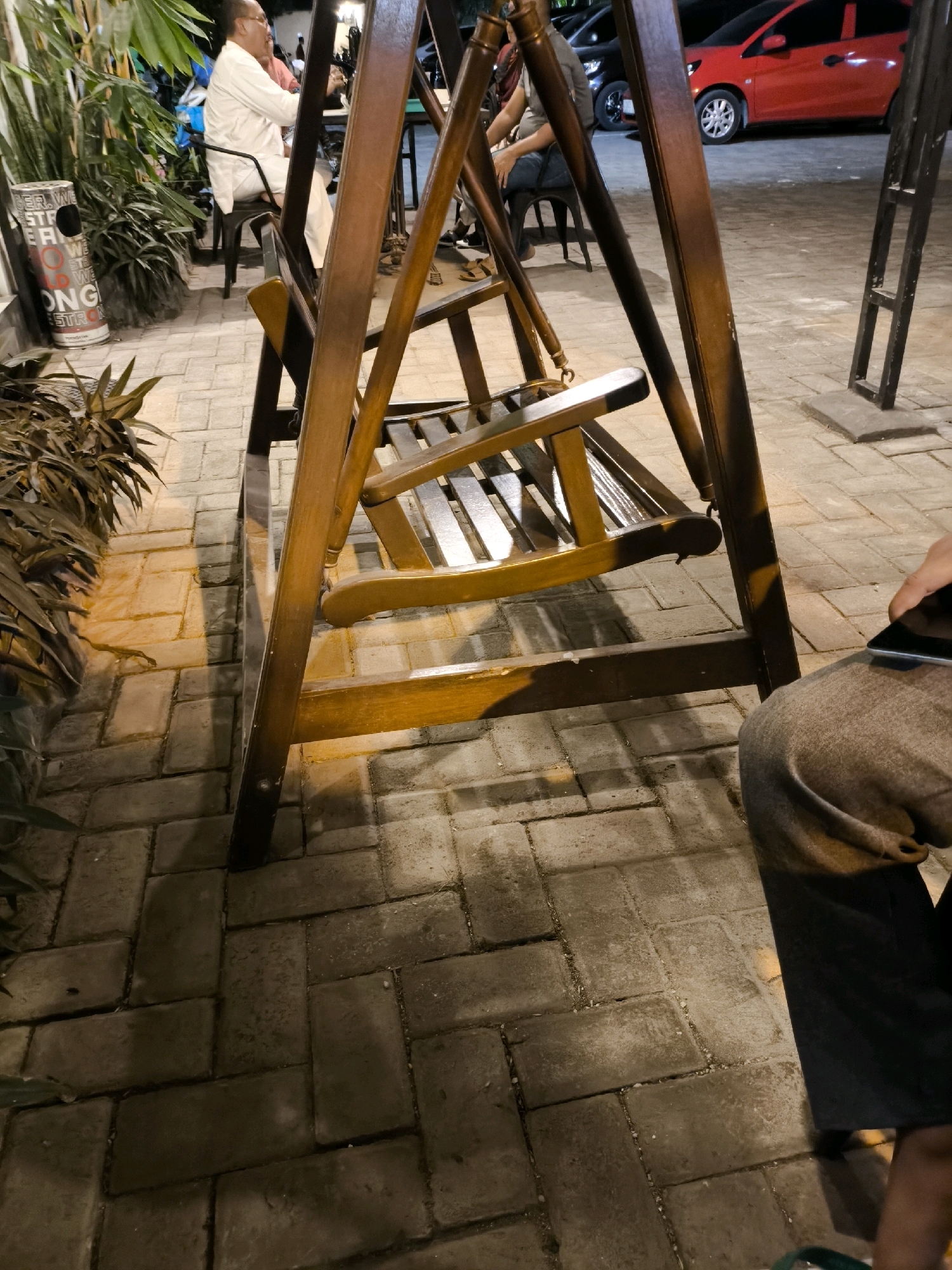 The image captures an outdoor seating area, likely a cafe or restaurant, at night. A wooden swing set is prominently featured in the foreground, with its seats partially obscured. To the left, a man in a white outfit is seated, and behind him, a red car is visible parked in the background. The ground is paved with rectangular interlocking bricks. A trash bin with text partially visible is also present to the left. The scene suggests a relaxed evening setting in a city environment, possibly Tuban, Indonesia, given the provided context, with artificial lighting casting shadows on the brick pavement.