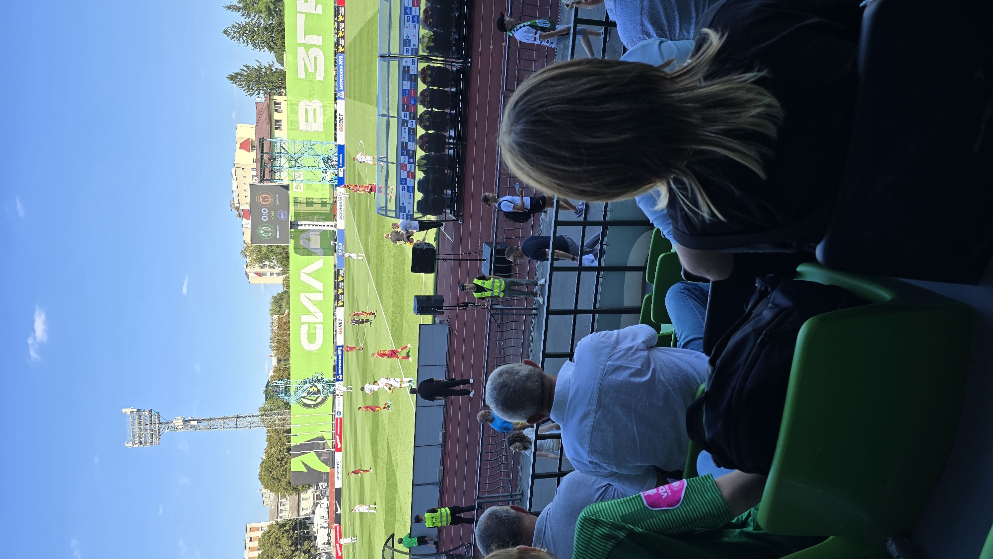 The image depicts a view from the stands of a football stadium in Zhytomyr, Ukraine. The scene is set on a sunny day with a clear blue sky. In the foreground, several spectators are seated, with their backs mostly to the camera, observing a football match in progress. The focus is on the playing field, which is a vibrant green turf.
A large scoreboard is visible in the distance, displaying the score "0:0". There are several players on the field, dressed in red and white kits, engaged in the game. Behind the field, a large green banner with Cyrillic lettering is prominently displayed. Structures resembling buildings and trees are visible beyond the stadium's perimeter. A tall floodlight tower stands on the left side of the image, a common feature of sports venues. The atmosphere suggests a live sporting event, with spectators present and the game underway.