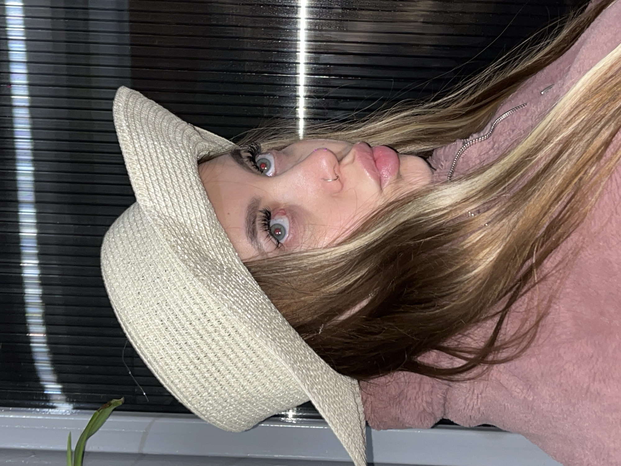 A young woman with blonde and brown hair wears a beige straw hat and a pink zip-up jacket. She is looking at the camera. The background consists of a black Venetian blind and a hint of green plant at the bottom left. There is no indication of the city of Novopokrovskaya, Russia.