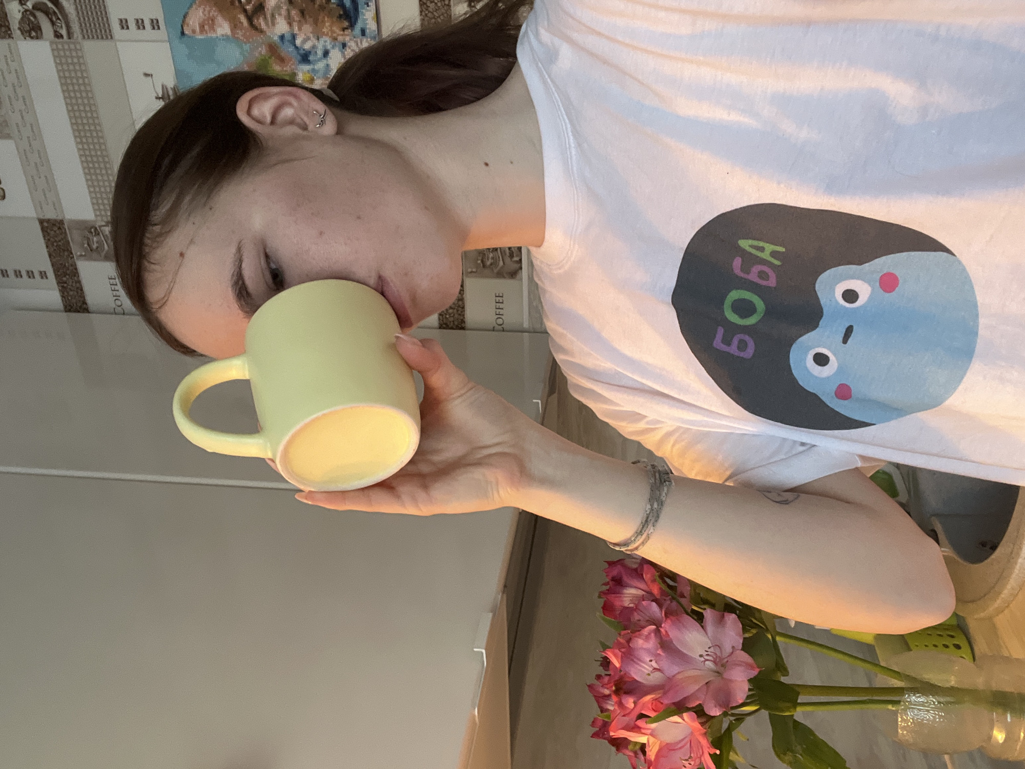 A young woman is drinking from a light yellow mug. She has dark hair pulled back in a ponytail and is wearing a white t-shirt with a cartoon graphic on it. She is holding the mug with her right hand, on which she wears a silver bracelet. A vase of pink flowers is visible in the lower right corner. The background includes a wall and what appears to be a shelf or countertop. There is no information to determine that the photo was taken in Chaikovsky, Russia.