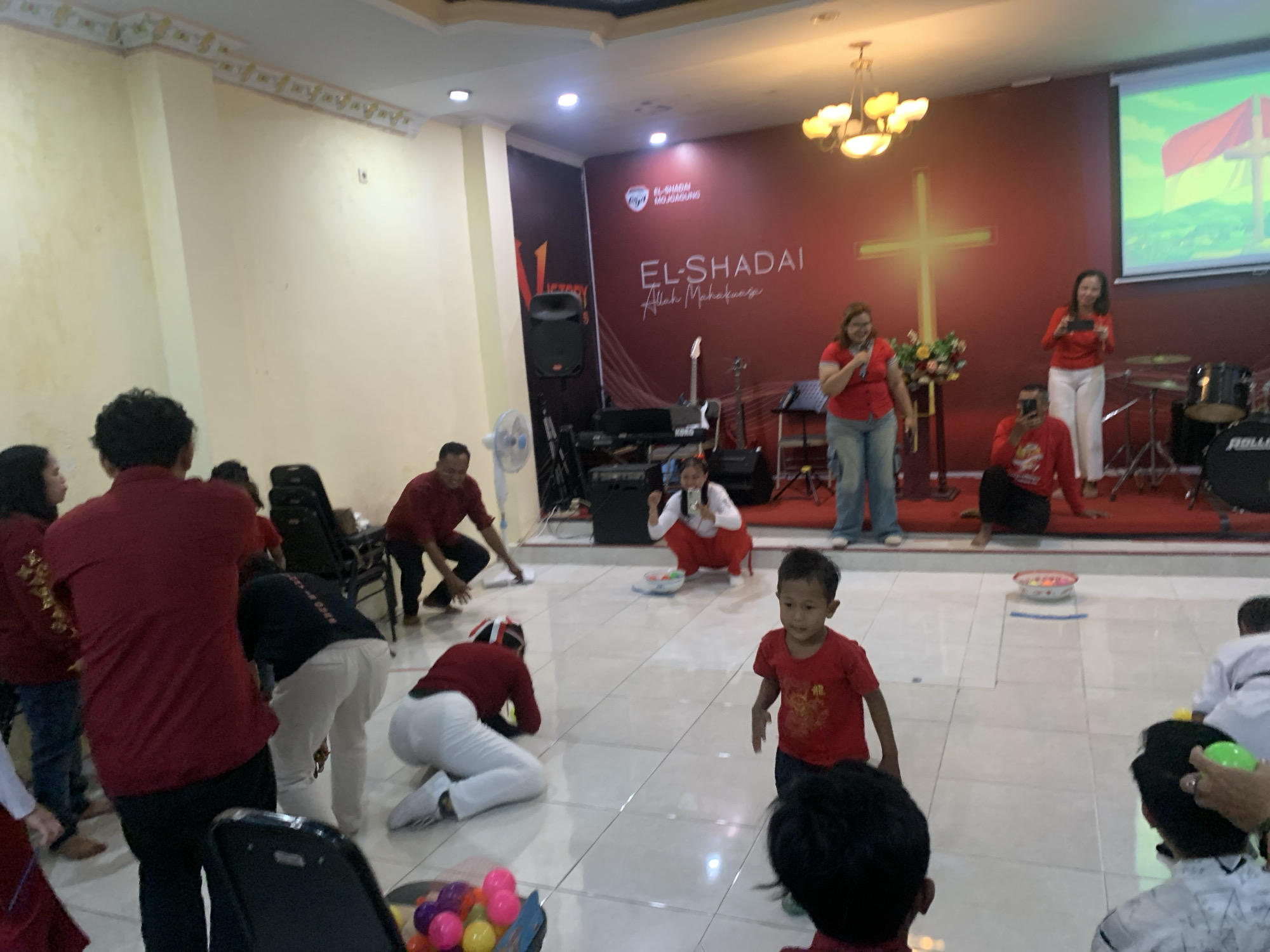 The image depicts an indoor event, likely a church gathering or celebration, taking place in Dukuh Mojo, Indonesia. The room has a stage area with a large red banner featuring the text "EL-SHADAI Allah Mahakuasa" and a cross graphic. A projector screen displays an Indonesian flag. Several individuals, predominantly dressed in red shirts and white pants, are engaged in activities on the tiled floor. In the foreground, a young boy in a red t-shirt stands looking towards the camera. Other people are seen sitting, kneeling, and interacting, suggesting a communal activity or game. Musical instruments, including a keyboard and drums, are visible on the stage, indicating a possible musical performance or worship service component. The lighting suggests an indoor artificial light setting.