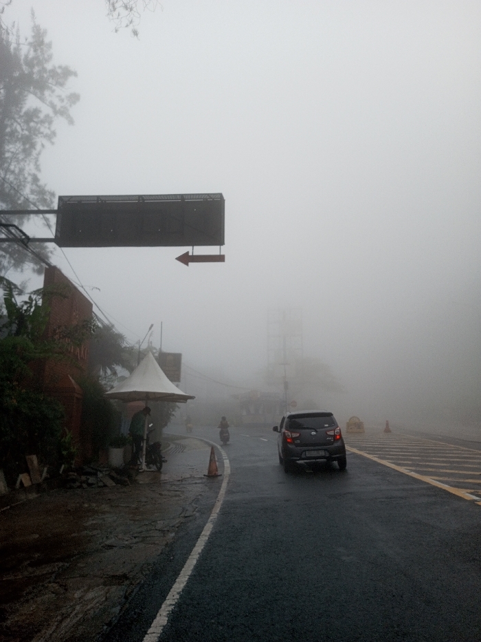 A car drives on a wet road through dense fog. A sign with a right-pointing arrow hangs above the road. A small business with an umbrella stands to the left. Two people are standing under the umbrella. A motorcyclist is also visible on the road ahead. Visibility is extremely limited due to the fog, obscuring background details like buildings or distant objects.
