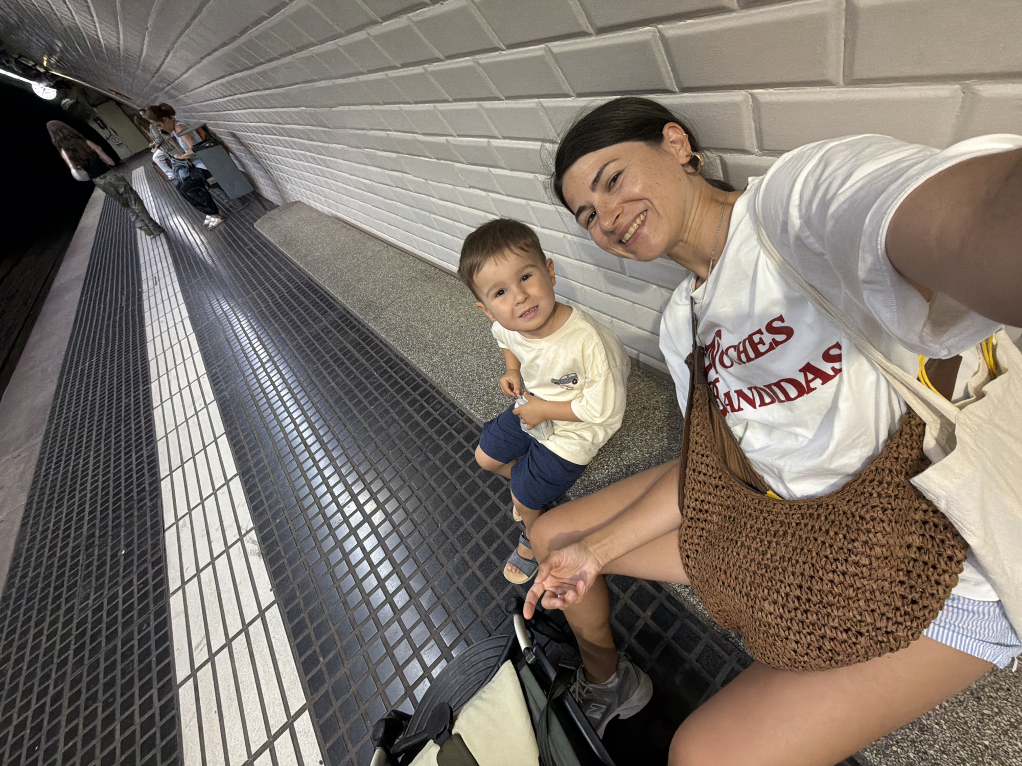 A woman and a young child are sitting on the ground next to a stroller on a tiled surface with black and white diamond patterns, possibly in a public transit station in Barcelona, Spain. The woman is wearing a white t-shirt with red lettering that reads "LOVES ADIDAS" and blue and white striped shorts. She is smiling and looking at the camera. The child is wearing a light-colored t-shirt and navy blue shorts. He is also smiling and looking at the camera. In the background, other people are visible walking. The lighting suggests it is daytime or a well-lit indoor area.