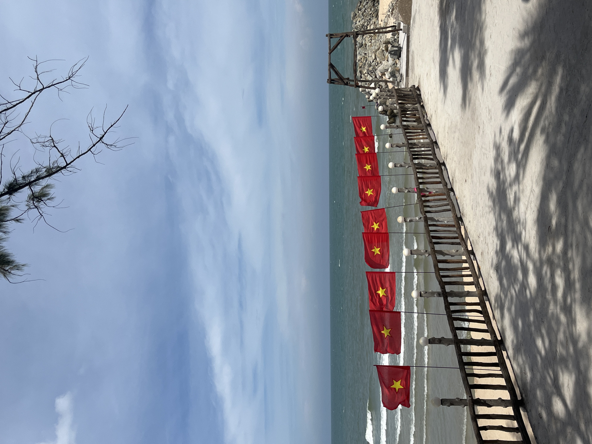 The image depicts a picturesque beach scene with the ocean and a cloudy blue sky. A row of red flags with yellow stars, which appear to be Vietnamese flags, are lined up along a wooden railing overlooking the water. The railing is supported by posts topped with lights. Beyond the railing, the sandy beach meets the choppy sea. In the background, a rustic wooden structure stands on a rocky area next to the beach, suggesting a park or recreational area. The foreground shows a sandy or concrete path with shadows of palm fronds cast upon it. The location is identified as Phu Thuy Ward, Vietnam. There are no visible people in the image.