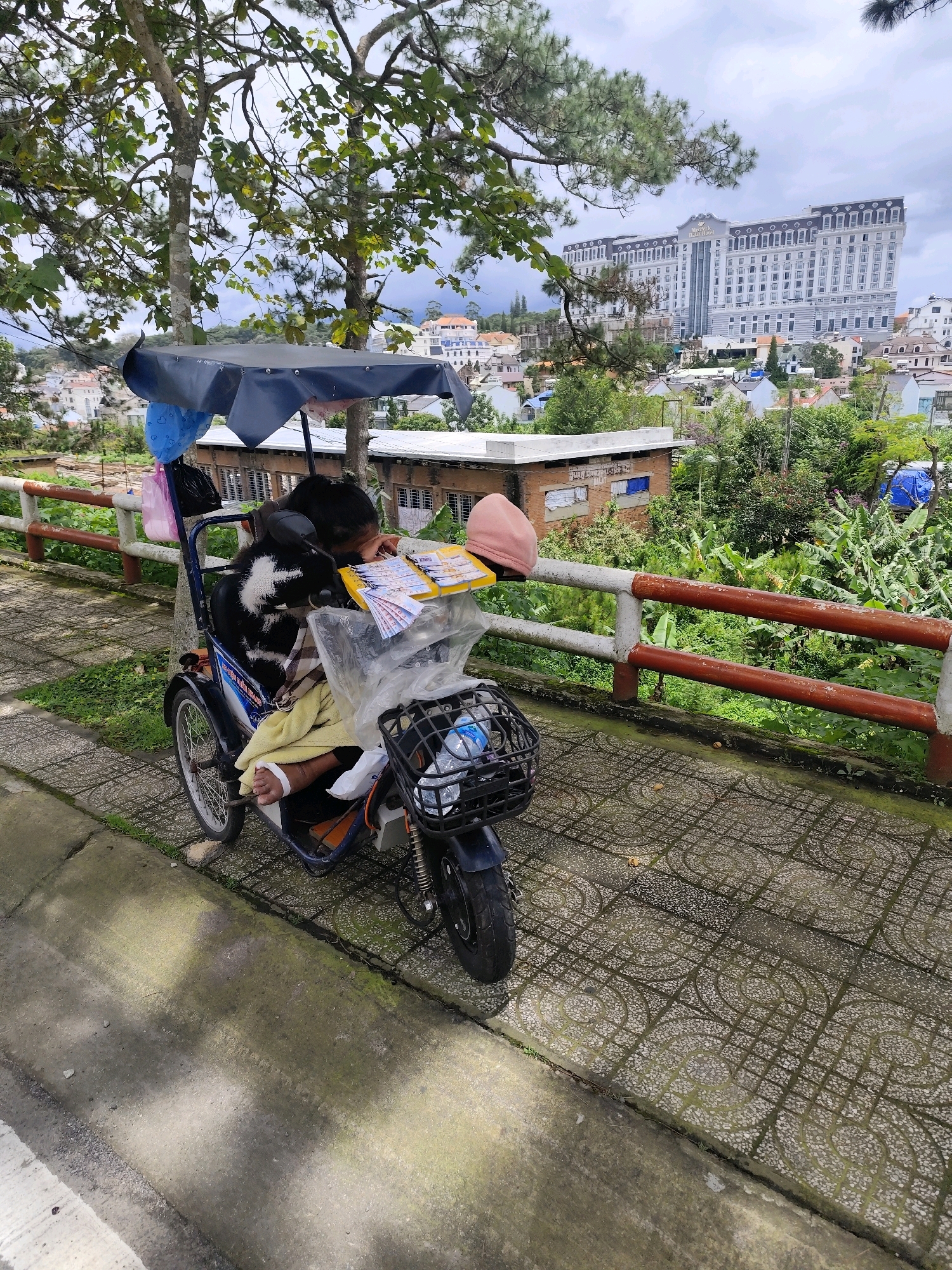 In Thành Phố Đà Lạt, Vietnam, a person is seated on a blue motorized scooter. The person is wearing a black and white patterned shirt and is leaning forward, appearing to be sleeping or resting. They are covered with a yellow blanket. A pink beanie is on their head. In front of the person, on the scooter's handlebars, is a yellow tray filled with what appears to be lottery tickets or similar paper slips. A clear plastic bag hangs from the scooter's canopy, and a black basket is attached to the front, containing a bottle of water. The scooter is parked on a sidewalk paved with decorative concrete tiles. A red and white striped railing runs along the edge of the sidewalk, overlooking a green, vegetated slope dotted with trees and buildings. In the background, a large, multi-story white building with a dark roof is visible, along with other smaller houses and vegetation, suggesting an urban or suburban setting within a hilly area. The sky is overcast.