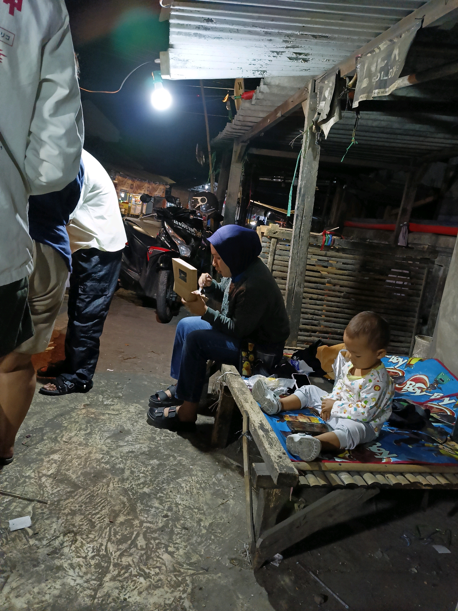 This image depicts a nighttime scene in Dompol, Indonesia. In the foreground, a young child is seated on a blue mat with cartoon characters. The child is wearing a patterned long-sleeved shirt and pants, and white shoes. To the left of the child, a woman wearing a blue hijab and dark clothing is seated and appears to be examining a small brown box. She is wearing dark sandals. To the left of the woman, two individuals are standing, one in a white jacket and the other in a white shirt and dark pants. Their legs and feet are visible, and they are wearing sandals. In the background, a motorcycle is parked. The environment appears to be an outdoor area with a corrugated metal roof overhead, supported by wooden structures. There is a visible light bulb illuminating the area. The ground is concrete and appears somewhat worn.