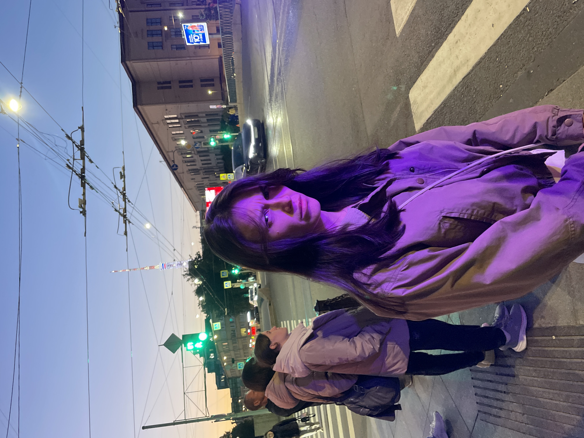 The image depicts an urban street scene at dusk in Saint Petersburg, Russia, as indicated by the prominent Saint Petersburg TV Tower in the background. The foreground features a young woman with dark hair, wearing a mauve jacket, looking directly at the camera with a slightly disgruntled expression. She is positioned near a pedestrian crossing, with white stripes painted on the wet asphalt road. To her left, a group of people are standing on the sidewalk, waiting to cross. Among them, a woman in a pink jacket and dark pants, and a man in a black jacket are visible. The street is illuminated by traffic lights, streetlights, and the headlights of a black car driving on the road. Overhead wires for trolleybuses crisscross the sky. Buildings with lit windows line the street, contributing to the city's nighttime ambiance. The overall lighting suggests a transition from day to night, with the sky displaying hues of blue and orange.