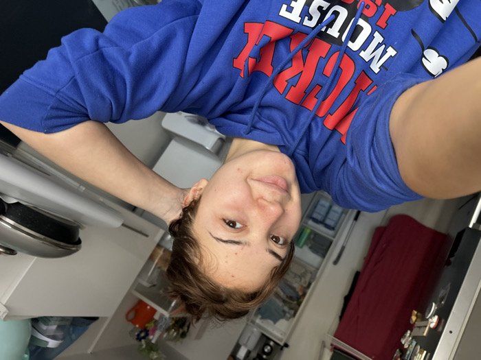 A frontal selfie captures a young adult individual, likely female, in an indoor residence in Schiltigheim, France. The individual has medium-length brown hair, pulled back, and is looking directly at the camera with a slight smile. They wear a royal blue hooded sweatshirt with "MICKEY MOUSE 1928" printed in red and white lettering, and a Mickey Mouse head graphic on the left sleeve. The individual's right arm is bent with the hand positioned behind their head. The background to the left features a metallic surface, possibly a refrigerator, with items affixed, a dark red garment hanging, and a white shelving unit containing clear plastic drawers with various items. The background to the right shows a white kitchen counter or cabinet with a silver pan and lid. Further back, a red pitcher and a vase containing artificial flowers are visible on a shelf. The scene is brightly lit.