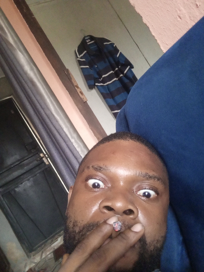 The image displays a close-up, upward-angled interior shot of an adult male, identified as being in Magboro, Nigeria. The man, possessing dark skin, a short beard, and short dark hair, occupies the lower portion of the frame. His eyes are wide open, revealing pronounced whites. He holds a lit cigarette or cigarillo in his right hand, positioned near his mouth, with a glowing red ember and ash clearly visible at its tip. A dark blue garment, likely worn by the man, is visible on the right side of the frame, near his head and shoulder.
The background consists of an indoor setting. A blue and white striped long-sleeved garment hangs on a hook against a light-colored wall in the upper-center. To the left, a dark-framed door or window is partially visible, adjacent to a dark grey curtain or drape. Two diagonal architectural trim pieces, one light peach and one dark brown, are also present on the wall, along with a small wooden fixture. The scene is illuminated by ambient, subdued lighting.