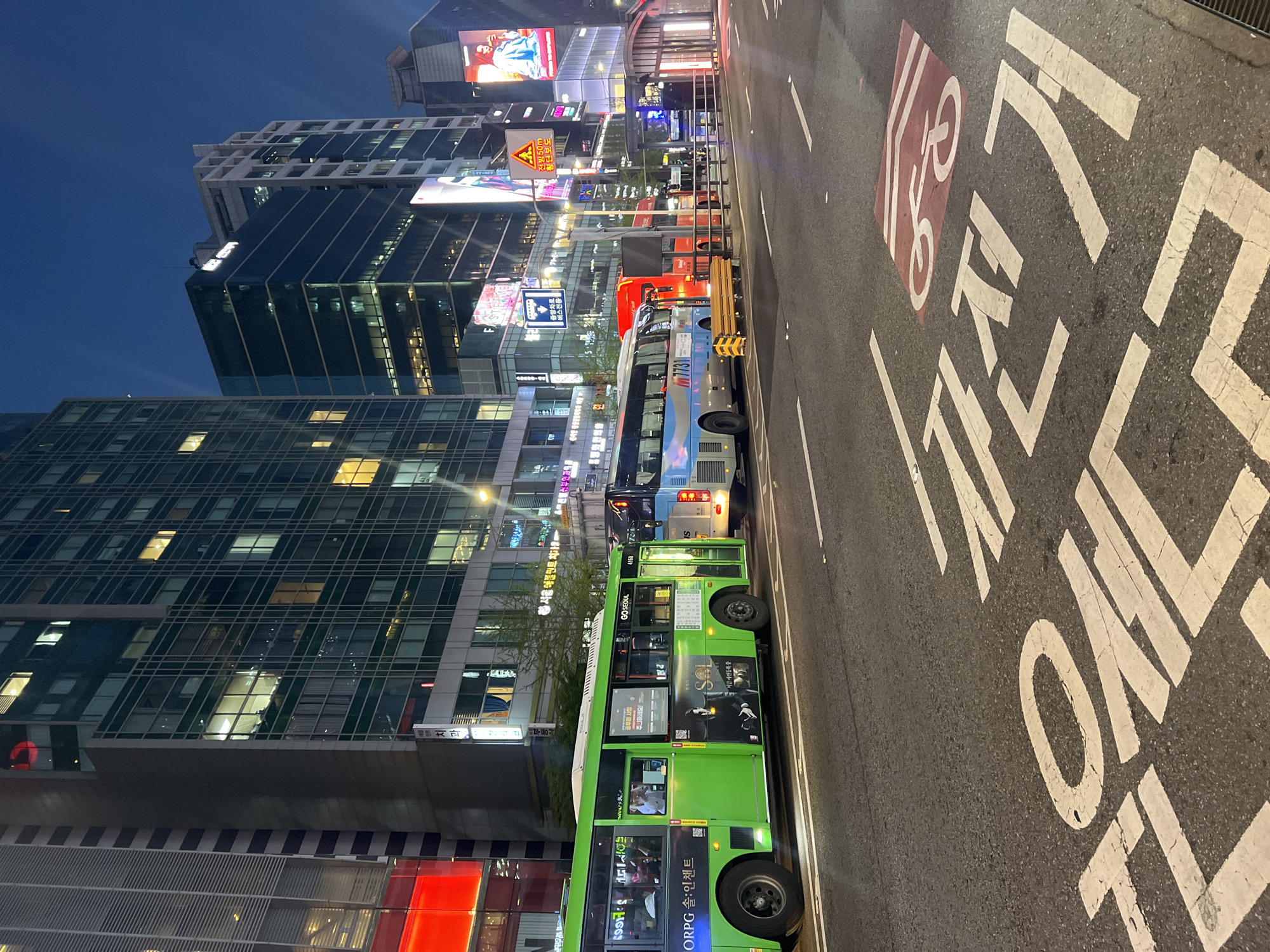 The image depicts a street scene in Seoul, South Korea, at dusk or early evening. Buildings line the street, some with illuminated windows and digital billboards. Several buses are parked along the roadside. The street is marked with white lines and lane markings, including a bicycle lane symbol and Korean characters that read "Central Bus Lane". Advertisement wraps are displayed on the sides of the buses. The scene is urban, with a mix of commercial and residential buildings. The light is low, suggesting it is late evening. There are signs written in Korean along the buildings and street signs.