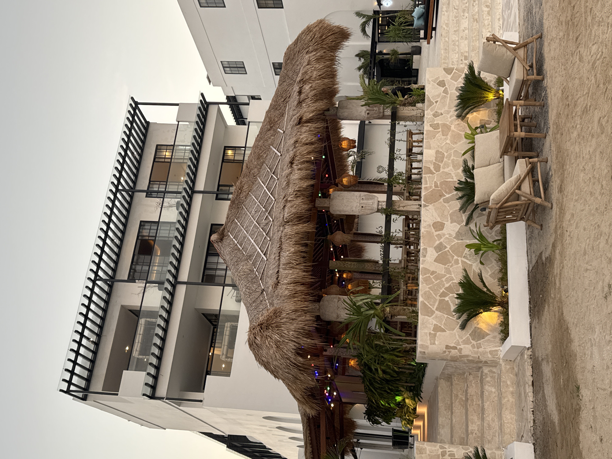 An outdoor scene features a multi-story white building with a thatched-roof structure in front of it. The thatched roof shelters a dining area with tables and chairs. String lights are visible hanging under the thatched roof. Several palm plants are situated around the structure and its stone base. To the right of the thatched-roof structure, a white building with dark windows is visible. In the foreground, a seating area with light-colored cushions and wooden furniture is placed on a sandy surface. Steps lead up to the thatched-roof area from the left side of the image. The sky above is overcast. This scene is located in Kuta Selatan, Indonesia.