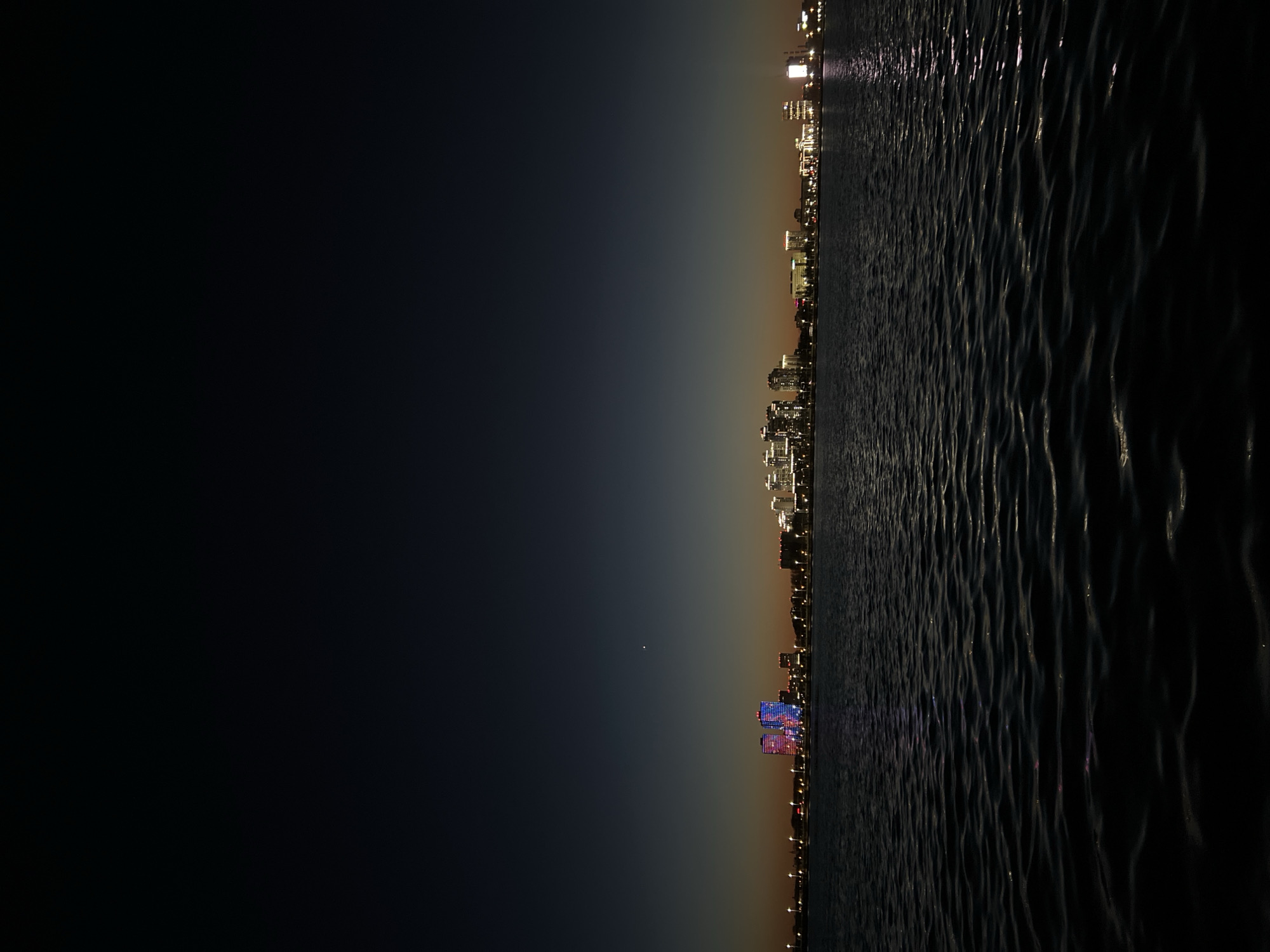 The image depicts a nighttime view of Kazan, Russia from across a body of water, likely the Volga or Kazanka River. The skyline is visible with building lights illuminating the cityscape. The water in the foreground shows ripples, possibly from a gentle breeze. The sky transitions from dark blue to a lighter gradient near the horizon where the city is situated. A single bright star or planet is visible above the city lights. Distinct, brightly lit buildings are seen toward the lower edge of the skyline, potentially indicating specific landmarks.