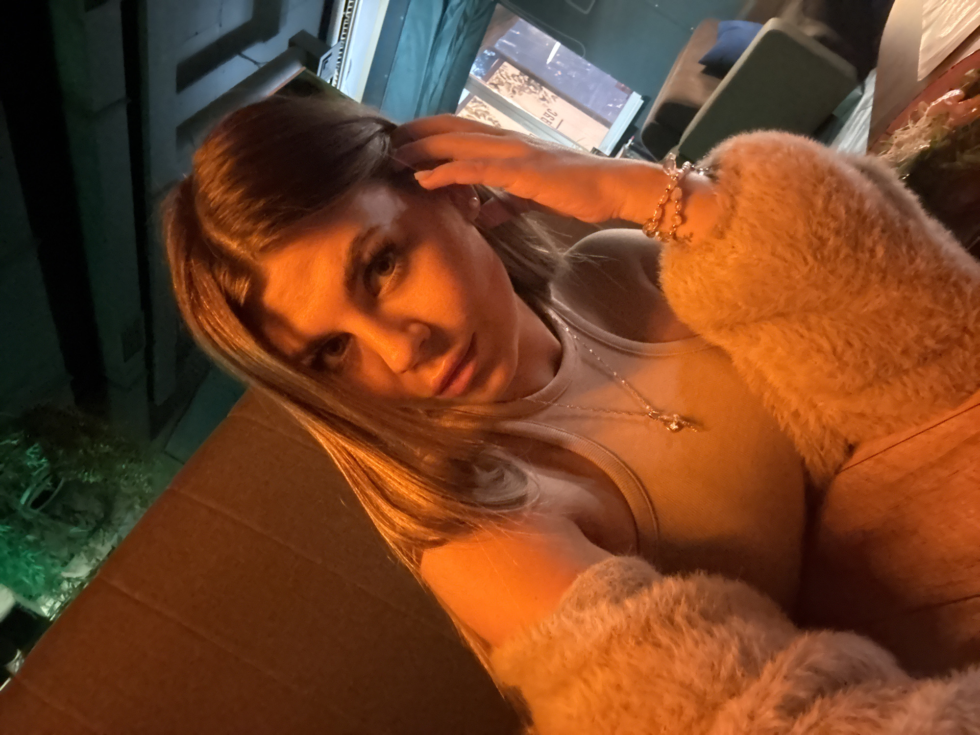 A woman is captured in a close-up selfie, her head and shoulders filling the frame. She has blonde hair, styled with some strands falling over her face, and is looking directly at the camera with a neutral expression. Her left hand is resting on her hair near her ear, and she is wearing a white tank top with a delicate necklace. Her right arm is covered by a fluffy, light-colored sleeve, and she is also wearing bracelets on her right wrist. The background appears to be an indoor setting with warm lighting, possibly a restaurant or bar. The environment is dimly lit with some industrial-style ceiling elements visible. The colors are dominated by warm tones of orange and brown from the lighting and surfaces, with some green accents in the background. The location in the city of Chaikovsky, Russia, cannot be determined from the visual information provided in the image.