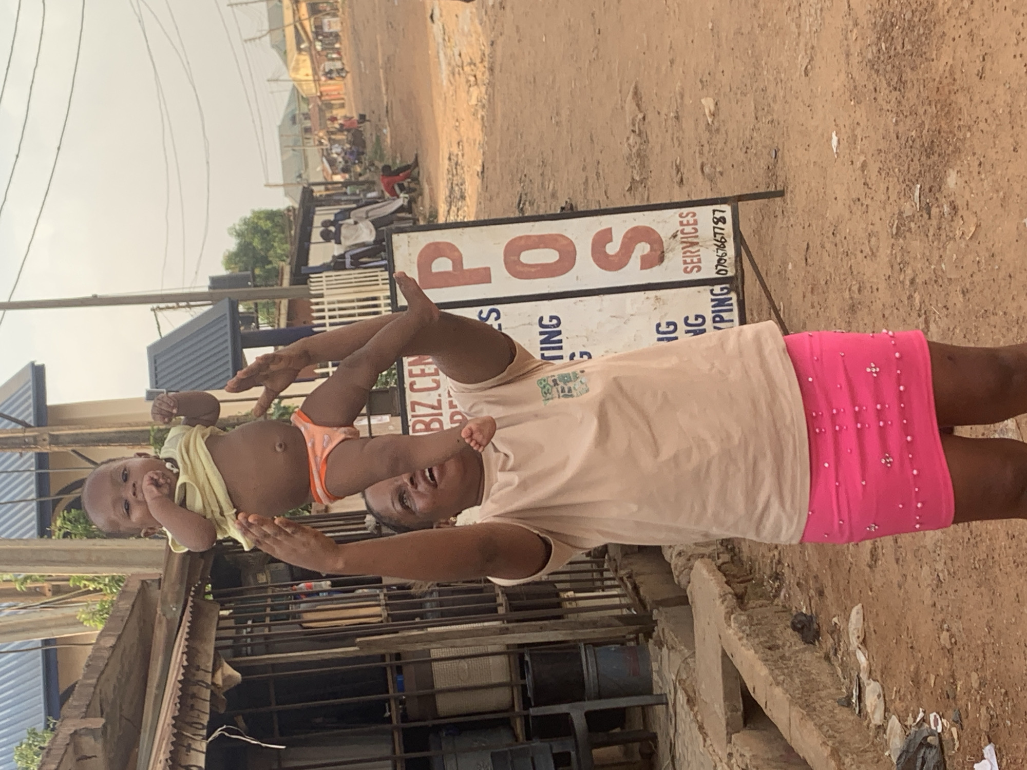 A woman in Sabon Tasha, Nigeria, wearing a peach-colored shirt and pink skirt, holds a baby up in the air with both hands. The baby wears a yellow shirt and orange underwear. Behind the woman is a sign for "POS SERVICES" with a phone number. The environment appears to be a street with buildings and people in the background. The ground is dirt.