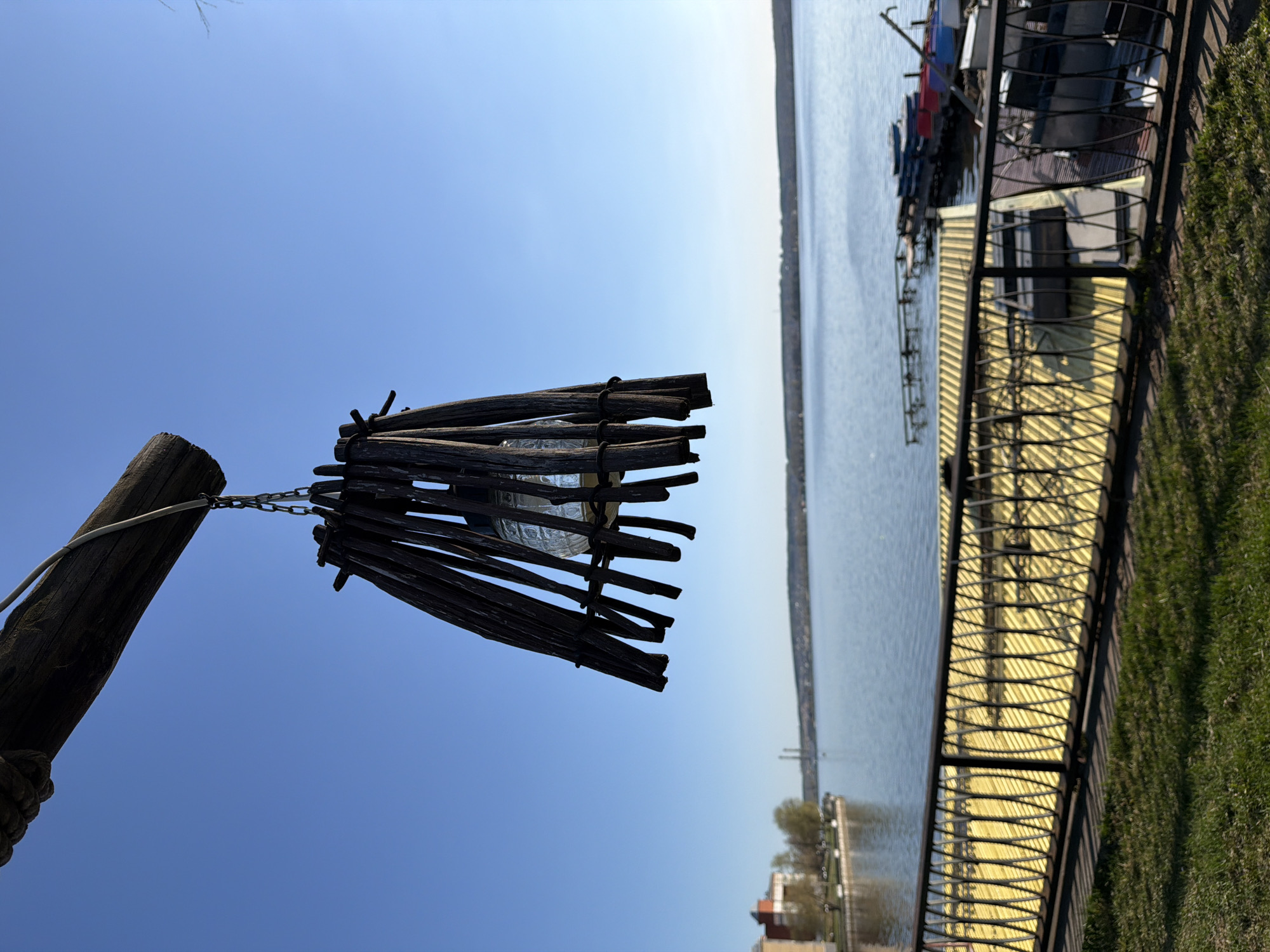 An outdoor lamp hangs from a pole by a chain, featuring a bulb encased in a wooden slat shade. Adjacent is a waterfront view of the Votkinsk Reservoir in Votkinsk, Russia. Boats are docked at a pier along the water's edge, which is edged by a fence and green grass. The sky is clear blue.