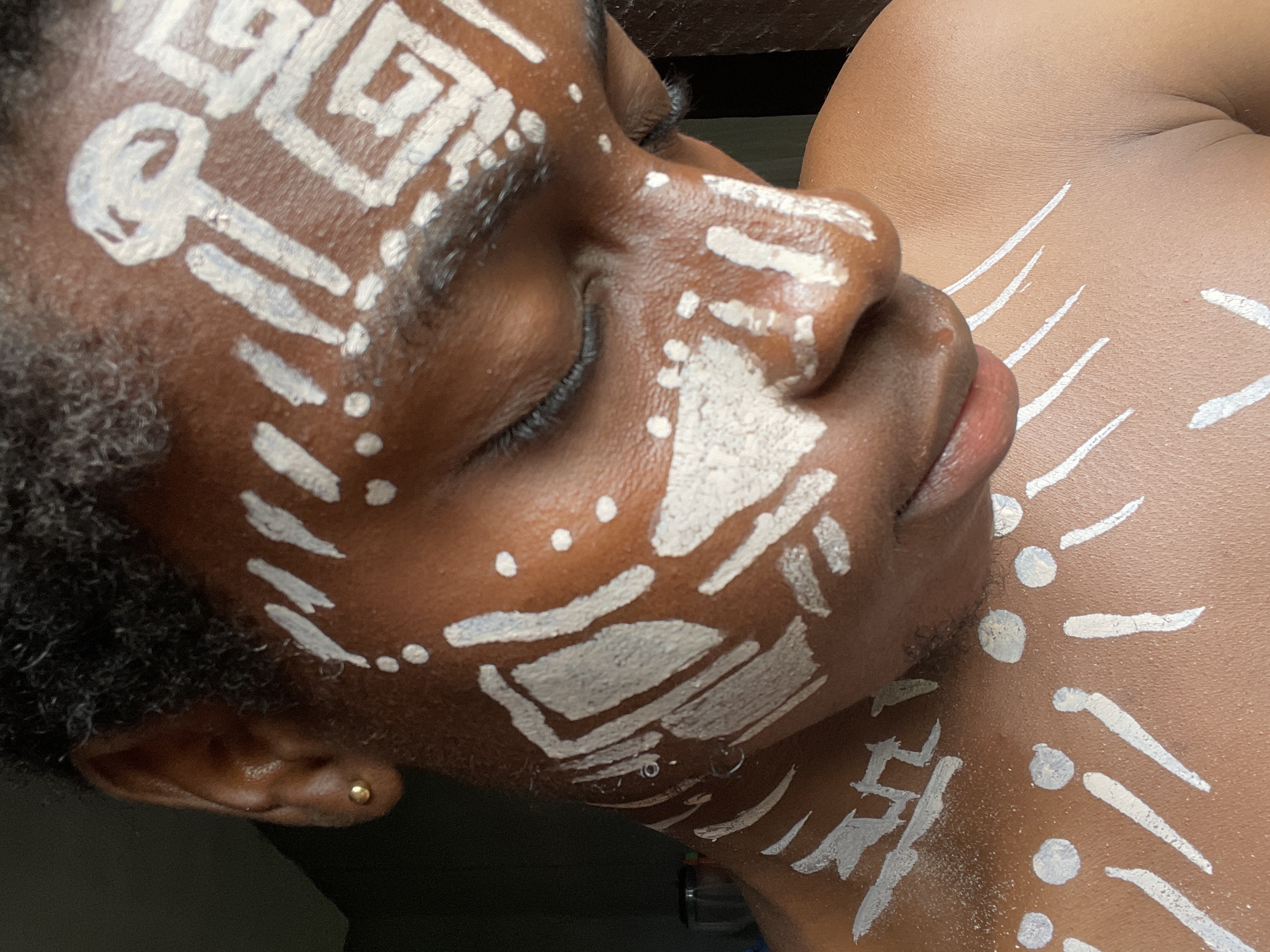The image features a person with dark skin lying down, adorned with white body paint. The paint is applied in geometric and tribal-like patterns on the face and chest. The individual's eyes are closed, and their expression appears peaceful. The background is dark and out of focus, drawing attention to the person's painted features. The style suggests a cultural or artistic expression, potentially related to traditional practices. There is no explicit evidence in the image to confirm this scene to be in the city of Onitsha, Nigeria.