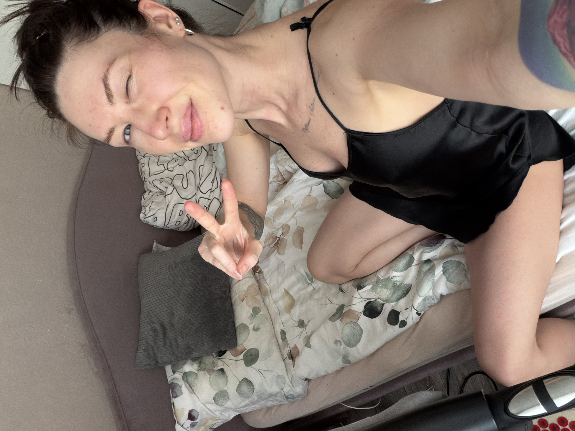 A woman is lying in a bed, making a peace sign with her left hand. She is winking. She is wearing a black silk top. The bed has a floral patterned comforter and several pillows. The scene is likely in an indoor location, possibly a residential home. There is no evidence in the image to confirm the location is in Perm, Russia.
