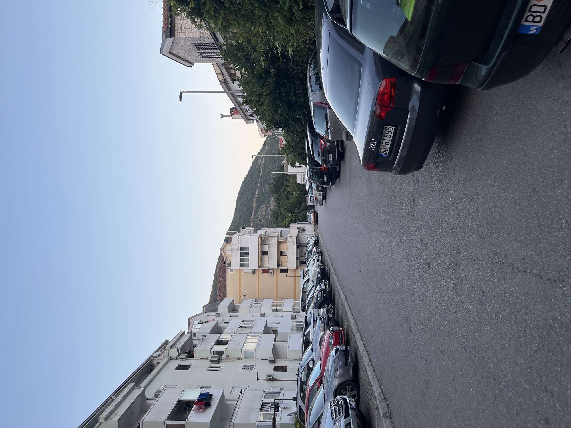 The image depicts a street scene in Budva, Montenegro. The street is lined with parked cars on both sides. On the left, there are apartment buildings with balconies, some of which have clothes hanging out to dry. The buildings are white and light yellow. On the right, there are more apartment buildings and trees. In the background, there are hills covered in vegetation. The sky is clear and light blue, suggesting it is either early morning or late afternoon. The overall scene is one of a residential street in a coastal town.