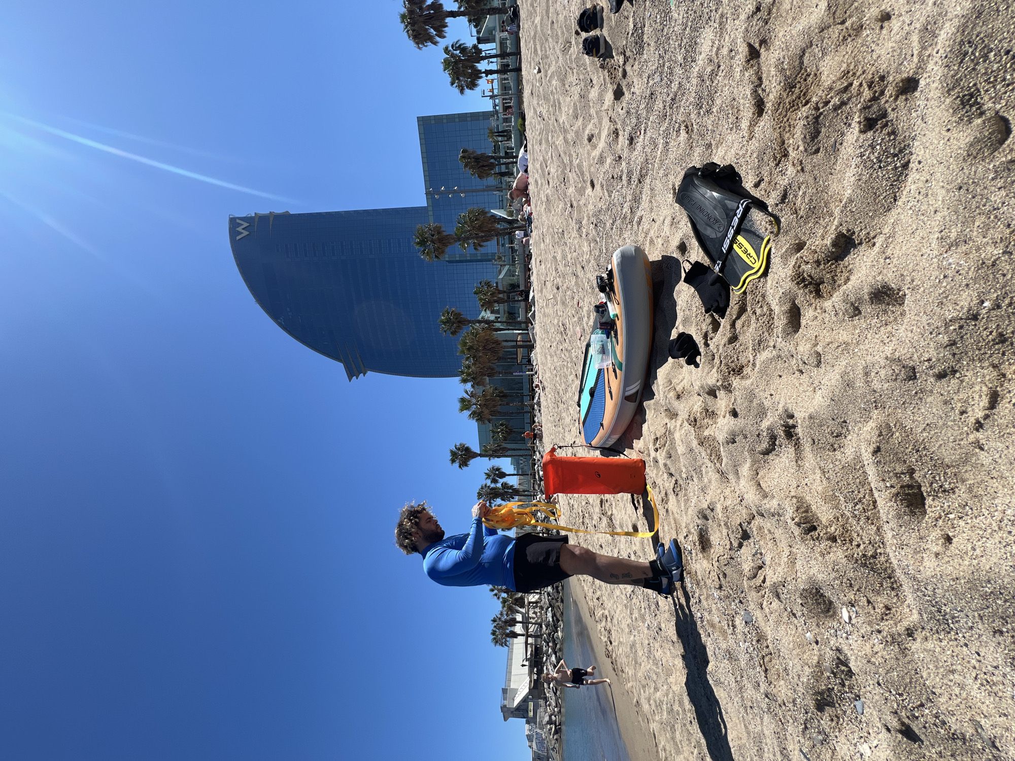 A daytime outdoor shot depicts a beach scene. A man with curly hair, wearing a blue long-sleeved shirt and black shorts, stands on the sandy beach. He is holding a yellow rope connected to an orange cylindrical bag. In the background is a distinctive curved building with the letter "W" on it, indicating this is likely the W Barcelona Hotel. Beside the bag is a paddleboard and diving fins on the sand. The sky is clear and blue. Palm trees line the edge of the beach. People are visible in the background on the beach and in the water.