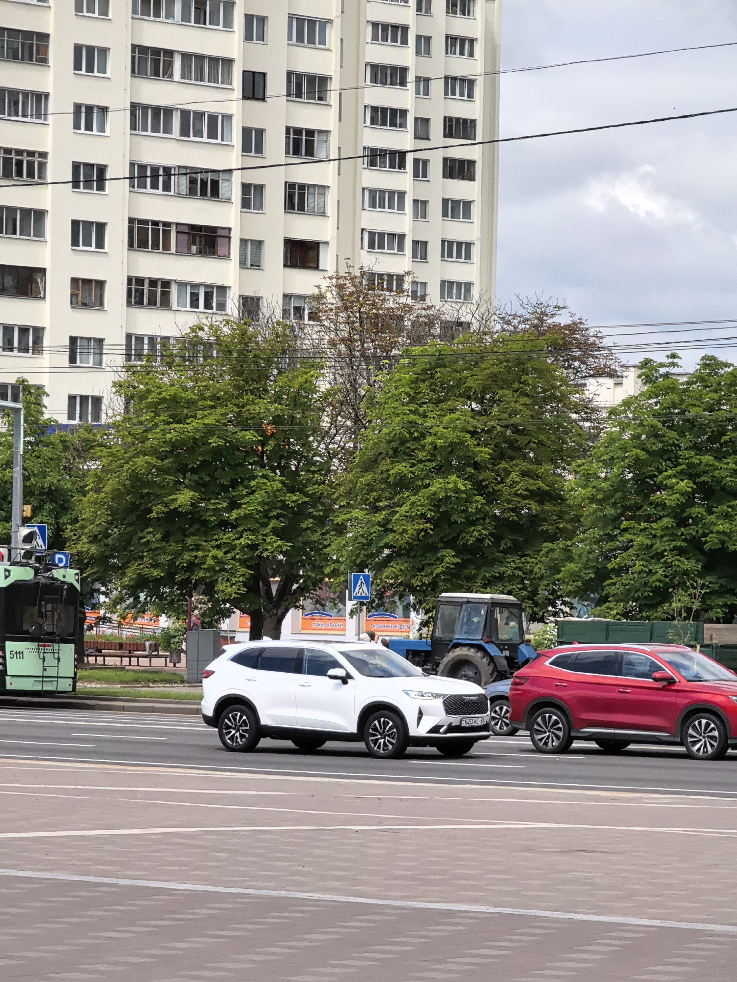 A street scene in an urban environment, likely in Eastern Europe, features a multi-story apartment building in the background with numerous windows and balconies. In the middle ground, a line of lush green trees partially obscures the building. A blue tractor is visible among the trees, and a green tram, numbered "5111," is stopped at the left side of the frame. In the foreground, two cars are driving on a paved road: a white SUV and a red SUV. The white SUV is in the left lane, and the red SUV is in the right lane, slightly behind it. Pedestrian crossing signs are present on the side of the road. The sky is overcast with white clouds.