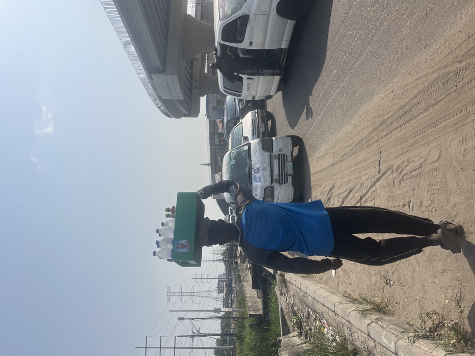 The scene depicts a street in Ikota, Nigeria, under a clear, bright sky. In the foreground, an individual, seen from behind, walks along a sandy dirt shoulder adjacent to a paved road. The person wears a blue and black long-sleeved shirt, dark pants, and sandals, and carries a green plastic crate on their head, containing multiple plastic water bottles and several smaller brown bottles. A small poster depicting a man's face is affixed to the front of the crate. The person's left arm is raised and bent at the elbow, hand near the crate.
To the left of the walking individual, the dirt shoulder is bordered by a concrete curb, beyond which lies an unkempt strip of ground with sparse vegetation and litter, leading to an area with electrical poles and large transmission towers in the distant background.
To the right, on the paved road, traffic is present. Immediately behind the foreground person, a white compact van, likely a commercial passenger vehicle, is visible, featuring text that includes "BEKE" and two small posters of a man's face on its side. Further down the road, another white van has its side door open, with a person standing at the entrance. Additional vehicles are observed in various lanes. Overhead, a concrete elevated road or overpass structure with railings curves across the upper right portion of the image. In the mid-ground, a slender, dark, spire-like structure rises from beyond the traffic. The overall lighting indicates strong sunlight, casting visible shadows.