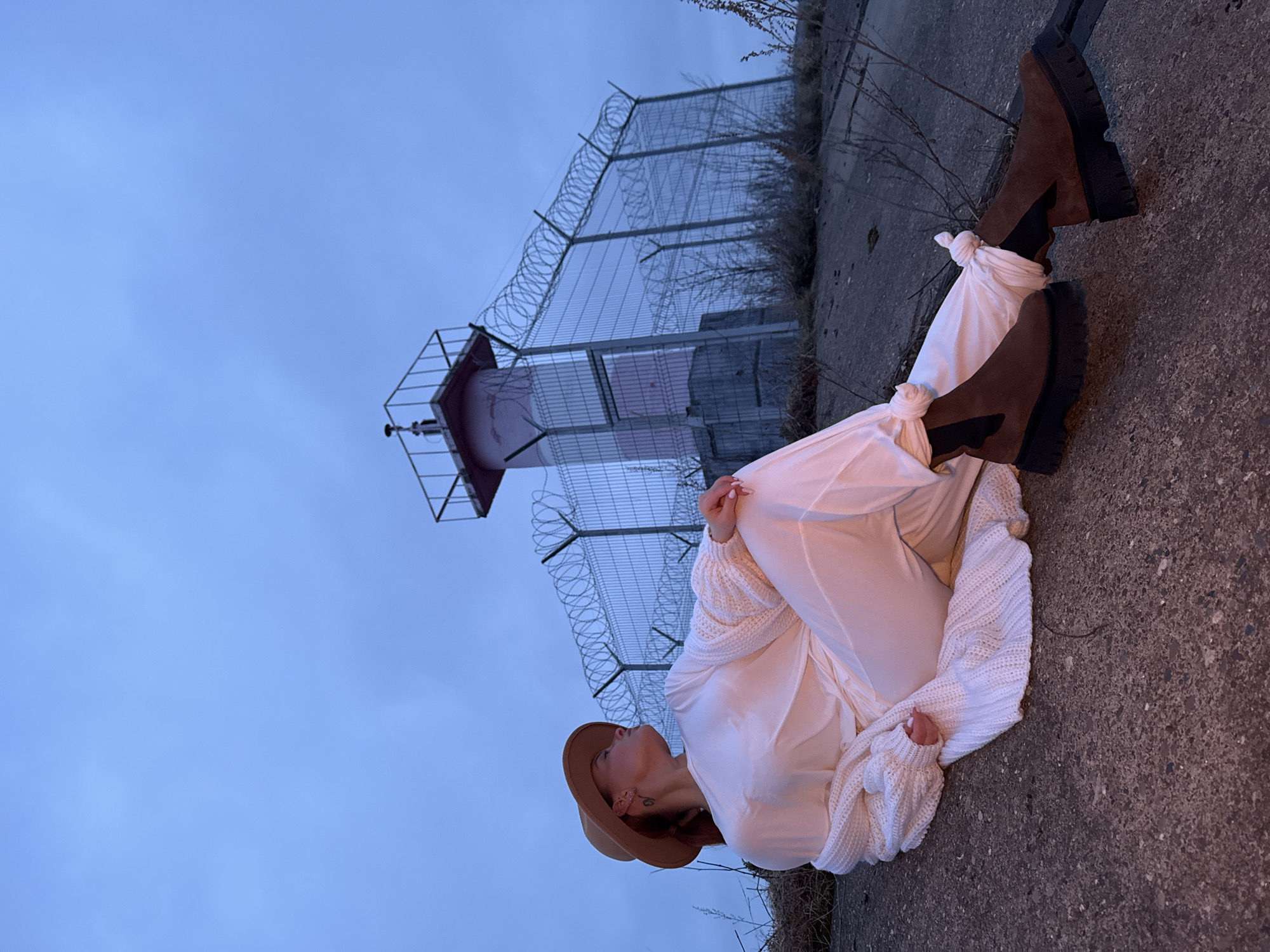 An image, rotated 90 degrees clockwise, captures a woman sitting on a rough concrete surface, looking upwards. She is positioned on the right side of the frame, wearing a broad-brimmed brown hat, a loose-fitting white shirt or dress, and a chunky white knitted cardigan. Her legs are extended, and her white garment is tied in knots around her ankles, revealing brown boots with thick black soles. Her hands are clasped in her lap, and her gaze is directed towards the sky.
In the background, to the left, stands a red-capped structure resembling a lighthouse or signal tower, enclosed by a tall chain-link fence topped with multiple strands of coiled barbed wire or razor wire. Dry, sparse vegetation grows along the concrete edge and behind the fence. The sky above is a uniform, overcast blue, suggesting twilight or an overcast day. The lighting is soft and muted, contributing to a serene or contemplative atmosphere. The scene, located in Chaikovsky, Russia, appears to be an industrial or utility area, possibly near a waterfront. No visible text is present in the image.