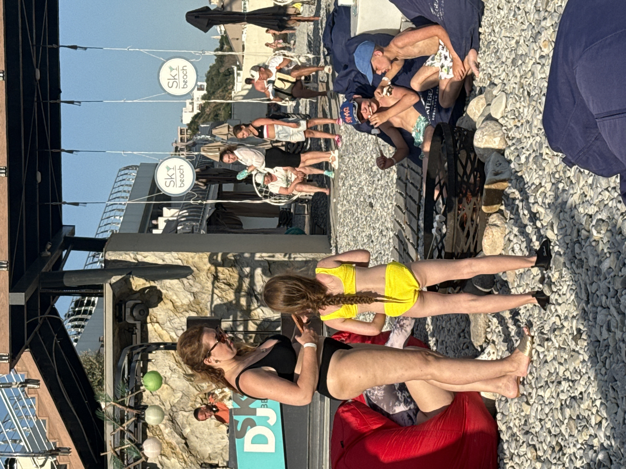 The image depicts a lively scene at what appears to be a beach club or resort, possibly in Yalta, Ukraine, given the provided context. Several people are visible, enjoying a sunny day outdoors. In the foreground, two women are seated on red beanbag chairs. One woman, wearing a black bikini, is looking towards the left, and the other, a young girl in a yellow bikini, is facing away from the camera. Behind them, more people are scattered on a gravelly surface, some reclining on blue blankets.
Further in the background, a row of white signs with circular logos and text that reads "Sky beach" are suspended from wires. Beyond these signs, buildings and lush green hills are visible, suggesting a coastal location with mountainous terrain. The overall atmosphere seems relaxed and casual, consistent with a vacation setting.
The time of day appears to be afternoon, indicated by the strong sunlight and shadows cast by the people and structures. The weather is clear and sunny, perfect for outdoor activities. The presence of beachwear and the "Sky beach" signs strongly point to a beachside or poolside establishment.
A metal fire pit is visible in the middle ground, adding an element of ambiance. The ground is covered in a mixture of gravel and paving stones. Among the people, some are dressed in swimwear, while others wear casual summer clothing. The overall impression is one of leisure and enjoyment on a warm day.