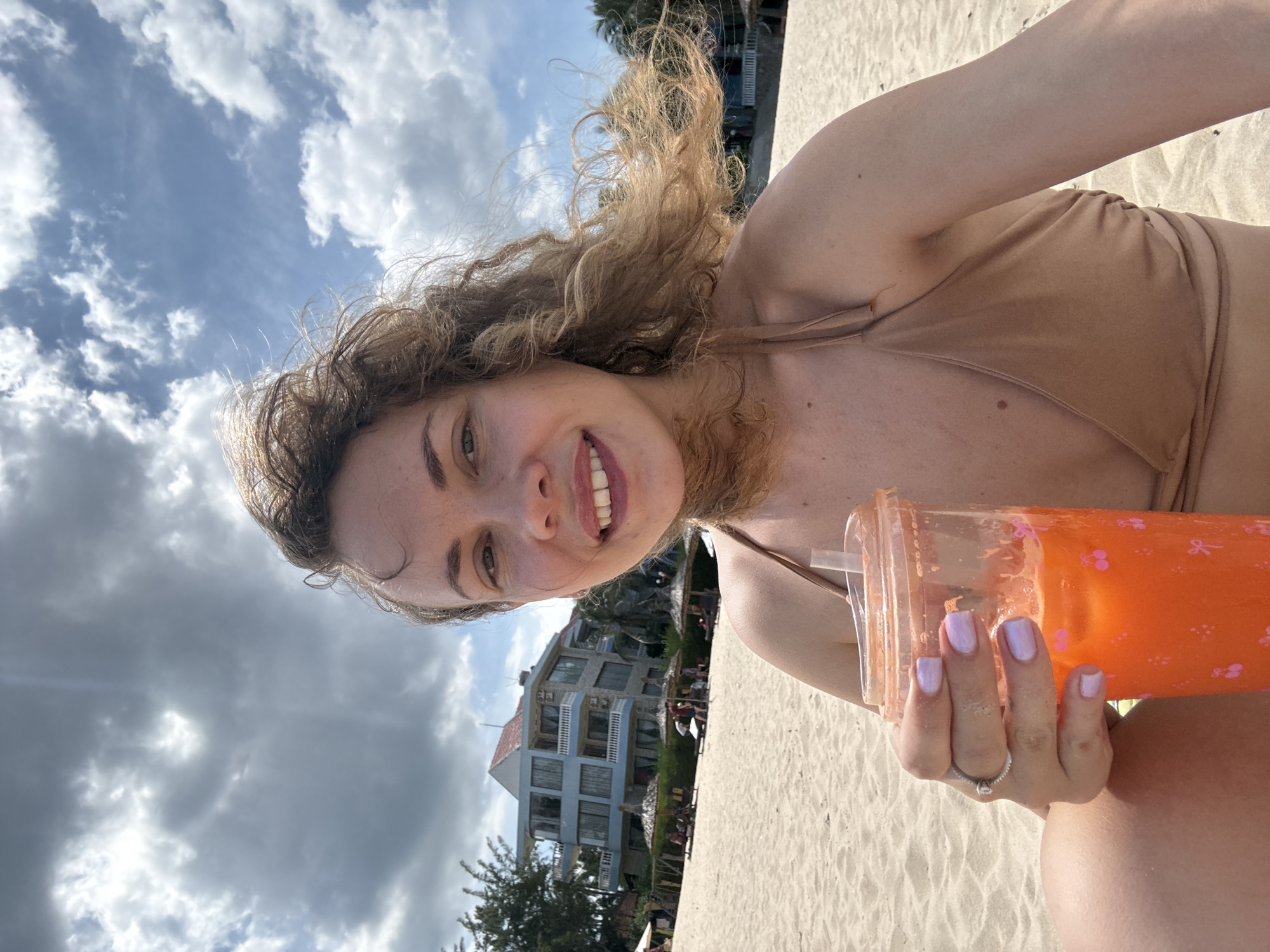A woman with curly brown hair is taking a selfie on a sandy beach in Phan Thiết, Vietnam. She is holding a clear plastic cup filled with an orange beverage and has on a light brown bikini. In the background, there is a building with multiple stories and balconies, as well as beach umbrellas and lounge chairs. The sky is cloudy with patches of blue.