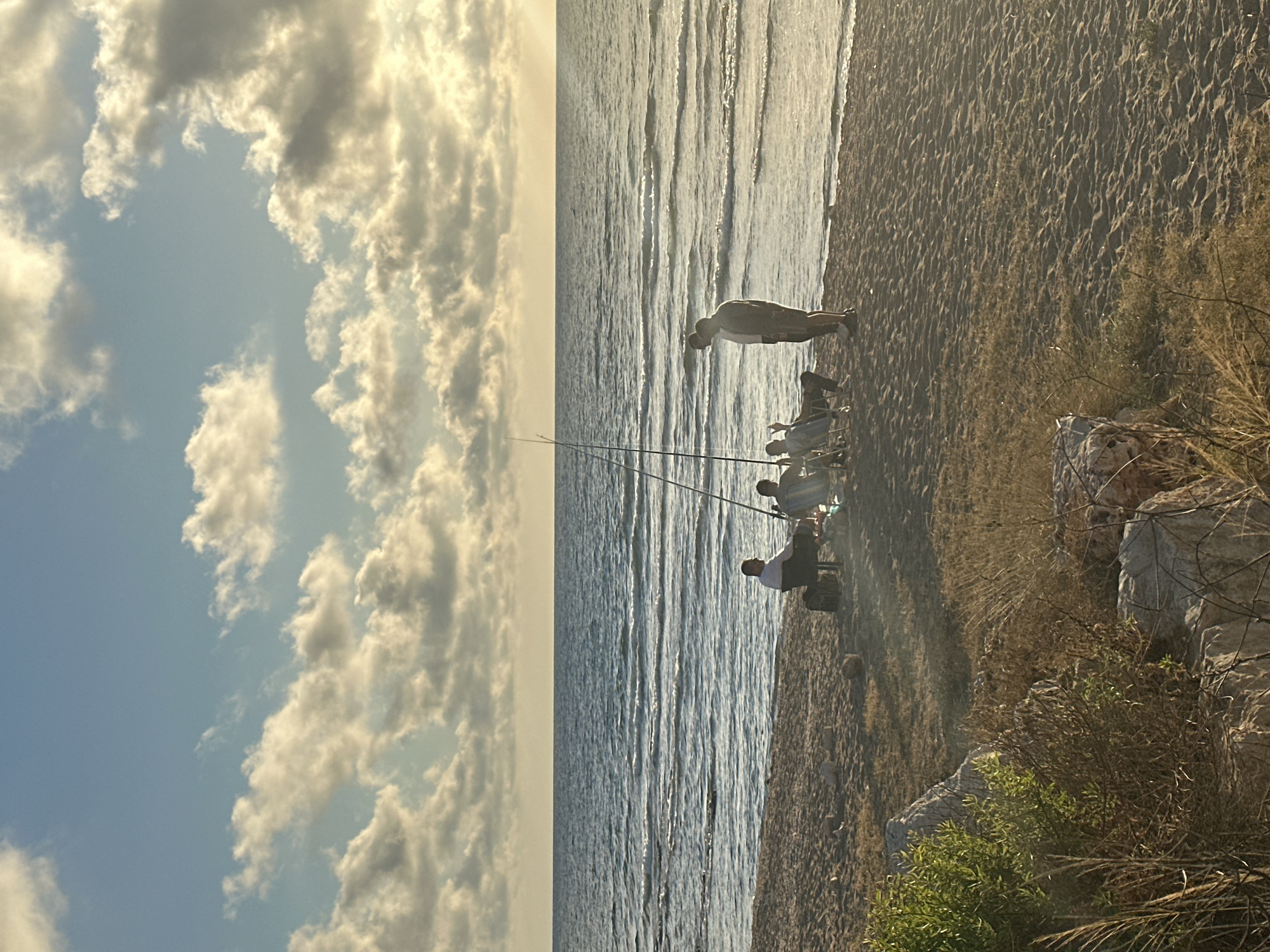 This media file captures a serene scene on a beach in Haifa, Israel, likely during the late afternoon or early evening, given the soft, golden light illuminating the sky and water. The sky is a prominent feature, filled with dramatic, textured clouds that stretch across the frame, tinged with yellow and white from the low-hanging sun. Below the sky lies the calm, rippling surface of the sea, reflecting the light and creating a shimmering effect.
The beach itself is composed of sand, with clear patterns of footprints visible, suggesting recent activity. The waterline marks a clear boundary between the sand and the water. Along the edge of the beach, dry grasses and some rocky outcrops are visible, adding a natural and slightly wild texture to the foreground.
Several people are engaged in fishing. One person stands near the water's edge, looking out towards the sea, while others are seated on chairs further back on the sand, holding fishing rods. The posture and arrangement of the people suggest a relaxed and patient pursuit of their hobby. Two fishing rods extend towards the water, emphasizing the activity.
The overall mood of the image is peaceful and tranquil, evoking a sense of calm and the enjoyment of a quiet moment by the sea. There are no visible signs or text in the image. The rotation of the image suggests it was captured in portrait mode and then rotated 90 degrees clockwise.