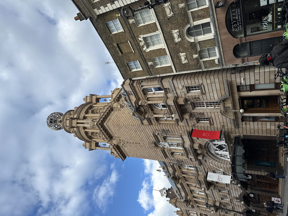 The image displays an urban street scene on St Martin's Lane in London, United Kingdom. The prominent structure on the left is the London Coliseum, an ornate, multi-story building constructed of light beige stone, featuring intricate architectural details, arched windows, and a grand entrance with a canopy. A tall, decorated tower rises from its center, topped by a large, metallic armillary sphere. A red vertical banner bearing "ENO" is affixed to the building's facade, alongside a white banner stating "Under 25s go Free." To the right, an adjacent building with a darker brick facade features white-framed windows and decorative moldings. Its ground floor is occupied by a commercial establishment named "Notes Coffee," with visible signage advertising "BRUNCH & SPECIALTY COFFEE" and "CRAFT BEER & COCKTAILS." A person is visible inside the coffee shop. On the pavement in the foreground, numerous shared bicycles are parked along the curb. Several pedestrians are present on the sidewalk, including one individual in the immediate foreground, with their back to the viewer, wearing a dark jacket and headphones. The sky above is partly cloudy with areas of clear blue visible.