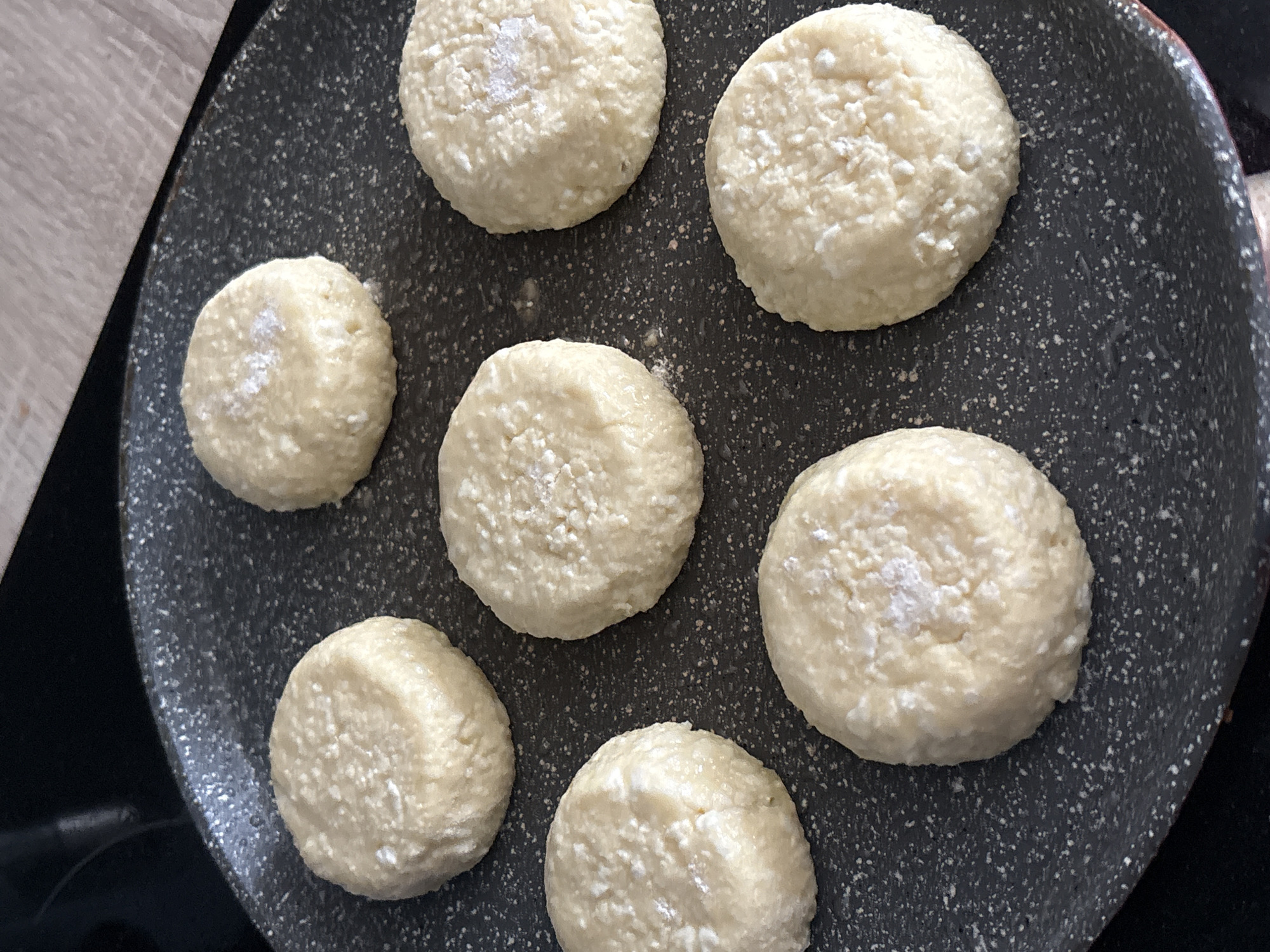 A top-down shot shows seven uncooked сырники (syrniki) on a gray speckled frying pan. The сырники are round, somewhat irregular, and appear to have a slightly textured surface, possibly due to cottage cheese, a key ingredient in сырники. Some of the сырники have a dusting of white powder, likely flour or sugar. The pan is speckled with white and gray dots. Part of a light-colored wooden surface is visible in the top left corner.
The location is not identifiable as the city of Chasovnya, Russia from this image.