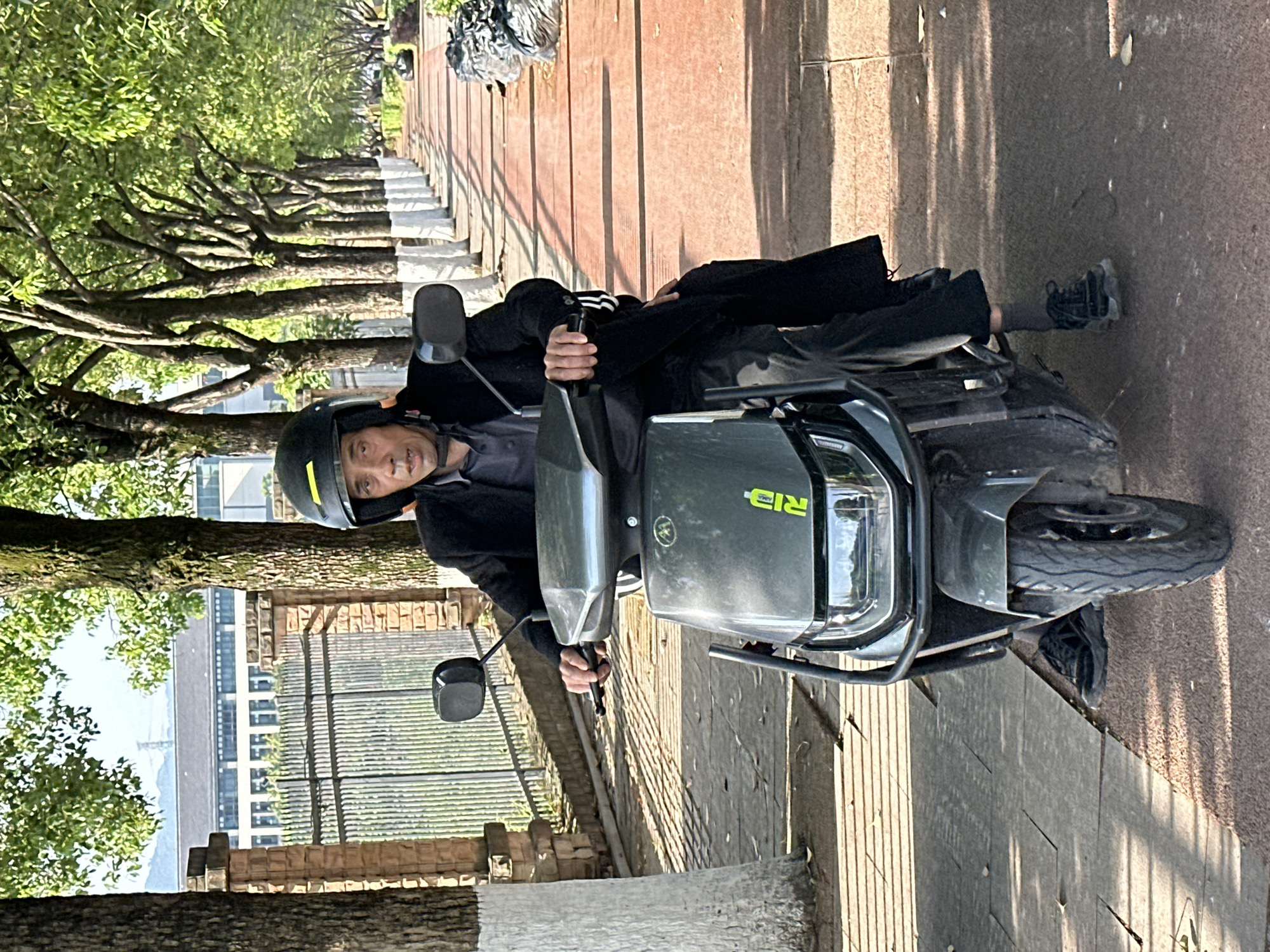An outdoor, eye-level shot captures an adult male sitting on a scooter on a sunny day in Jiangjin, China. The man, wearing a black helmet with a yellow accent, a dark long-sleeved shirt, and dark trousers, is positioned centrally on the scooter, looking directly at the camera with a neutral expression. His hands are on the handlebars. He wears black sneakers and dark socks, with his feet resting on the ground, indicating the scooter is stationary.
The scooter itself is dark grey or black, with "RID" in green stylized text visible on its front panel, along with a circular yellow logo. The foreground shows a path composed of reddish-brown paving stones and darker grey concrete sections, bordered by a line of mature trees with lush green foliage. These trees line a wide path, creating an avenue-like setting.
Strong shadows are cast on the path and ground, suggesting bright sunlight, likely mid-morning or afternoon. In the background, beyond the trees and a low brick wall, structures resembling multi-story buildings are visible, as well as distant hills or mountains under a clear sky. A pile of discarded material, possibly rubbish in a black bag, is visible further down the path on the right side. The overall scene depicts a moment of rest on a journey along a scenic, tree-lined route in an urban or semi-urban environment.