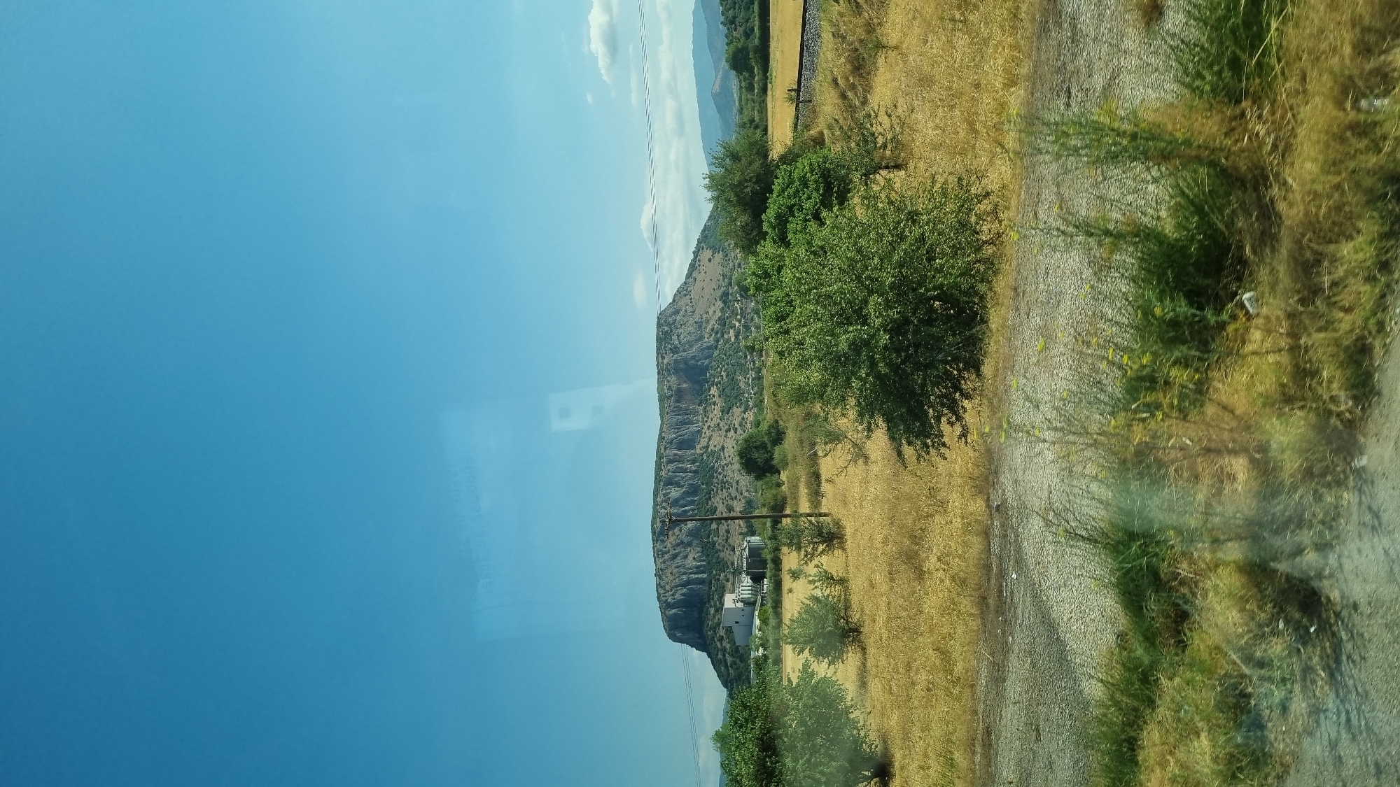 The photograph captures a scenic view of a large, flat-topped mountain in Theopetra, Greece. The mountain's rocky, tiered surface dominates the background, with a clear blue sky above. In the midground, a cluster of white buildings, possibly industrial or residential, is situated at the foot of the mountain. A solitary utility pole stands in front of the mountain, and the surrounding landscape is characterized by dry, yellow grass and scattered green trees. A dirt road or path runs through the foreground, bordered by overgrown vegetation. The scene appears to be a daytime landscape photograph, likely taken from a moving vehicle, as suggested by reflections on the glass surface in the upper portion of the image.