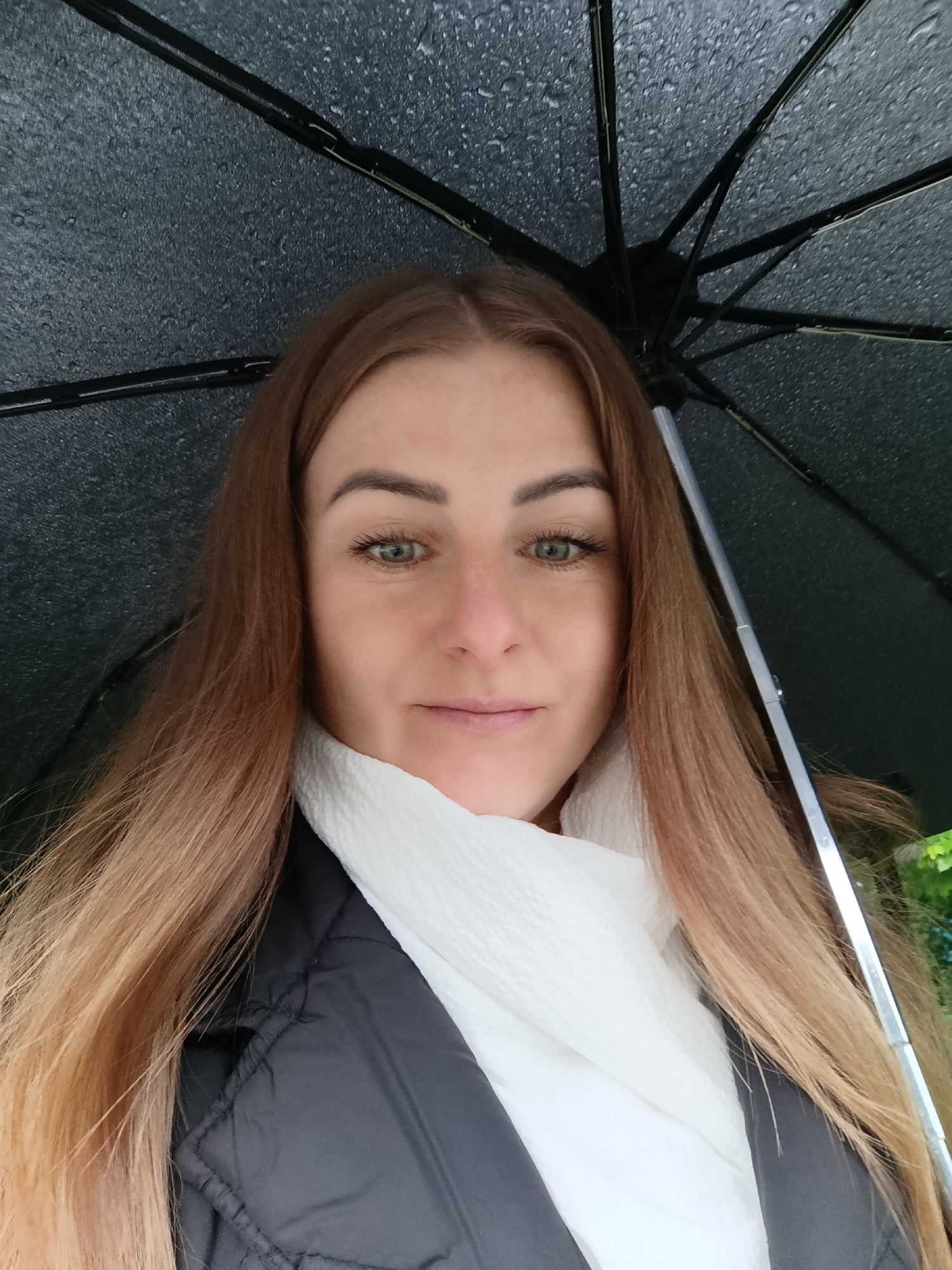 A woman is pictured in a close-up shot, holding a black umbrella. The umbrella is visibly wet with raindrops. She is wearing a white scarf and a black jacket. Her hair is long and ombre, transitioning from brown at the roots to blonde at the ends. The location is Unknown, Unknown.