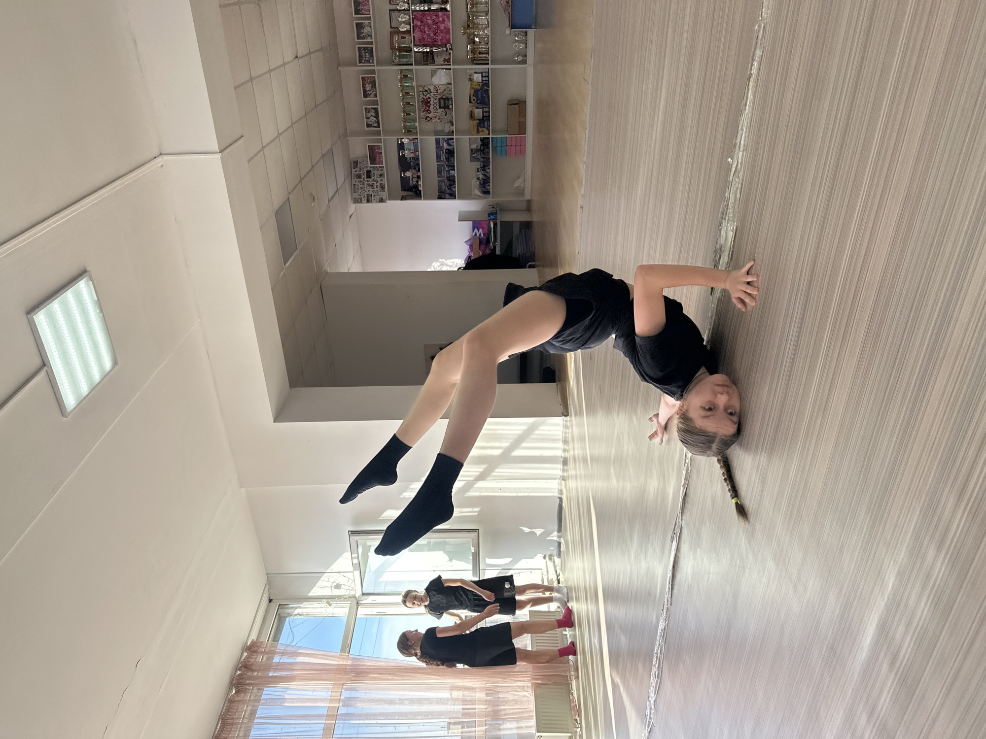The image depicts a young girl performing a headstand in what appears to be a dance studio. The studio has a light-colored wooden floor and white walls. The girl is wearing a black outfit and socks. Two other individuals, likely dancers, are visible in the background near a window covered with a sheer curtain. A mirrored wall reflects the scene, adding depth to the image. Shelves filled with trophies and other items line a wall further in the background. The location is possibly in Votkinsk, Russia, but there is no concrete evidence of such in the photo alone.