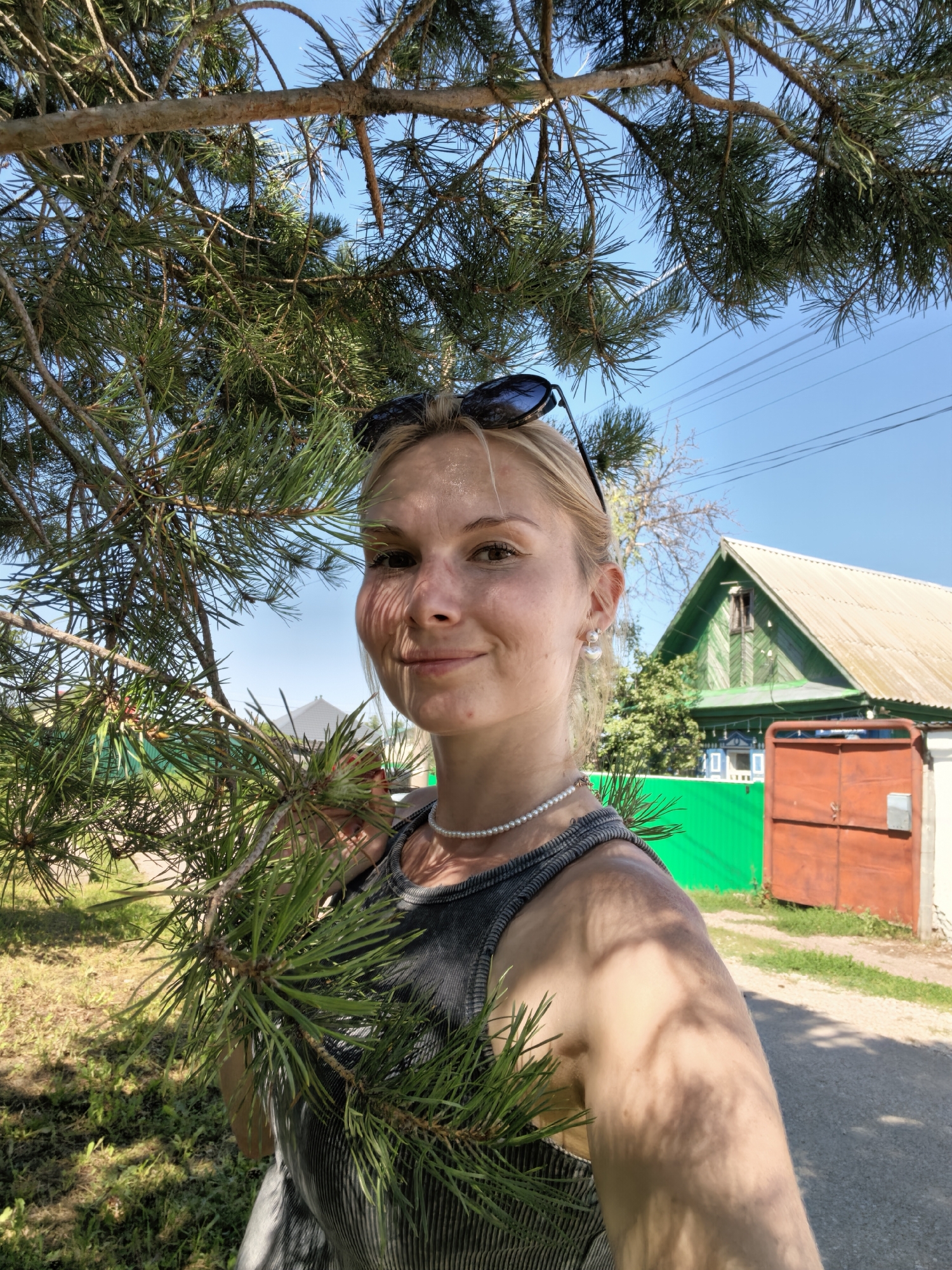 A young, fair-skinned woman with blonde hair and sunglasses on her head takes a selfie outdoors. She is wearing a gray tank top and a pearl necklace. Her right arm is visible, and there are shadows of pine needles cast on her skin. Behind her, a green wooden house with a metal roof is visible, along with a bright green fence and a large orange gate. The sky is clear and blue, and power lines are visible in the distance. The ground is covered in grass and dirt. The scene appears to be a residential area in Sterlitamak, Russia.