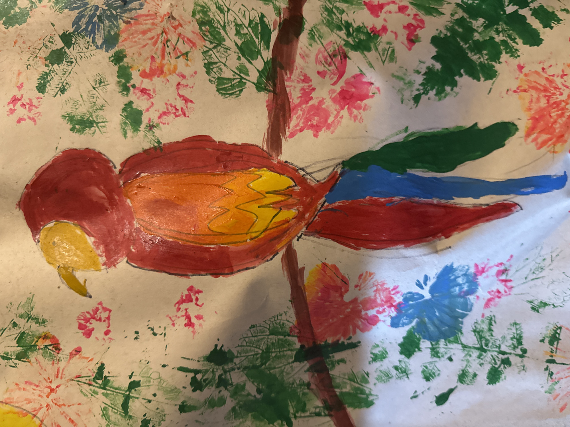 The image is a child's painting of a parrot perched on a tree branch amidst foliage and flowers. The parrot is rendered in red, orange, and yellow, with blue and green accents on its tail. The tree branch is brown. The surrounding foliage and flowers are depicted in various shades of green, pink, and blue using a stamping technique. The background is white. There is no location of Mèo Vạc Commune, Vietnam evident in the image.