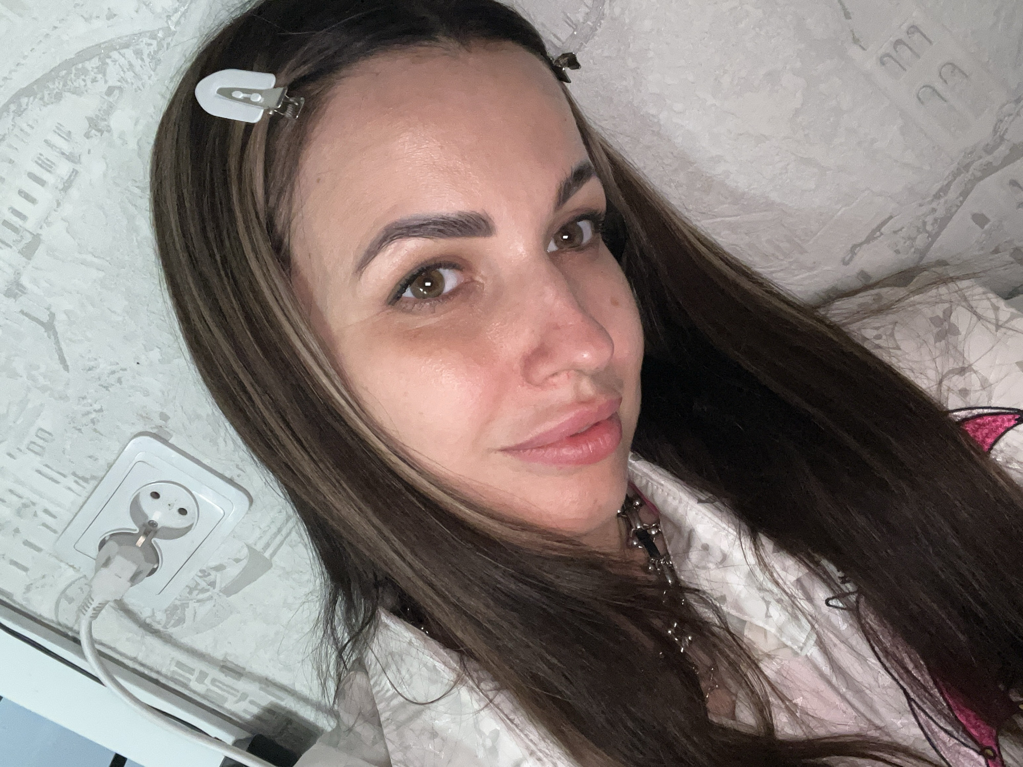 A woman with brown hair and green eyes is looking at the camera. She has two hair clips in her hair. She is wearing a white shirt. The background is a wall with a white electrical outlet and a plug. The city is Unknown, Unknown.