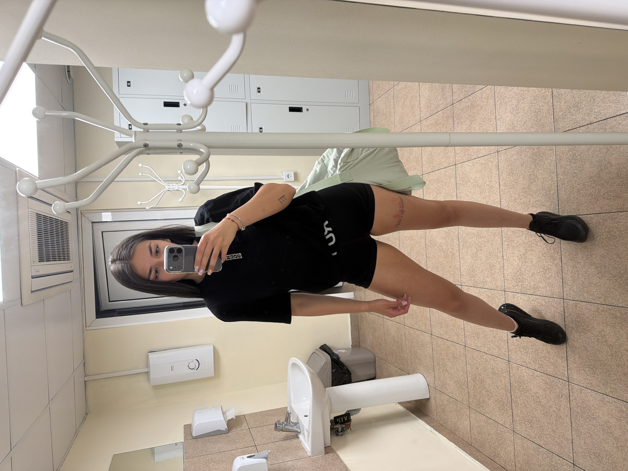 A young woman is taking a selfie in what appears to be a public bathroom. She is wearing a black t-shirt and shorts, and black sneakers. She is holding a phone in her right hand, using it to capture the image. The bathroom features tiled floors, a white sink and tap, and a wall-mounted soap dispenser. There is a rack for coats or bags. Behind her are lockers, suggesting this might be a sports facility or public area. The image was not captured in Gabrovo, Bulgaria.