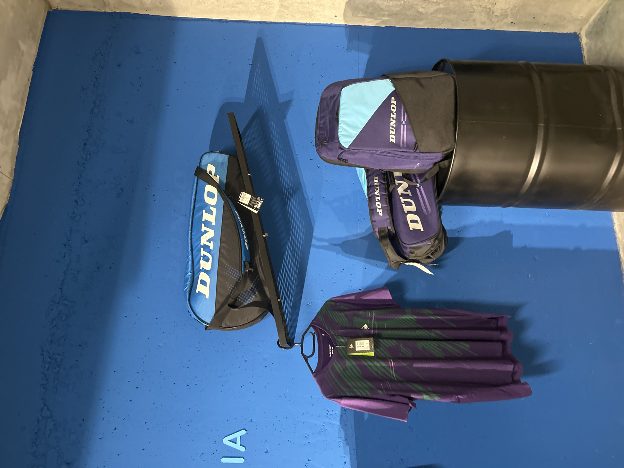 The image depicts a display of Dunlop brand sporting goods, specifically tennis bags and apparel, against a bright blue, textured wall. The wall has some visible imperfections and appears to be painted, possibly with a concrete or stucco finish. On a black metal shelf mounted on the wall, there is a blue and black duffel bag with the "DUNLOP" logo in white letters. Below this, a purple t-shirt with a green diagonal stripe pattern is hanging on a black hanger from a hook. The t-shirt has a brand tag attached. To the right of the t-shirt, a larger purple and blue backpack with the "DUNLOP" logo is displayed, with a black duffel bag placed in front of it. These bags are resting on top of a large black cylindrical barrel, which appears to be made of metal. The lighting casts shadows on the wall, suggesting an indoor environment. The phrase "IA" is visible in large, light blue block letters on the far left of the wall, though its context is unclear.
The provided information states that this scene is located in Tbilisi, Georgia. Without further information about the specific establishment, it is not possible to pinpoint the exact location within the city. However, the display suggests a retail environment, possibly a sports equipment store or a dedicated Dunlop outlet.