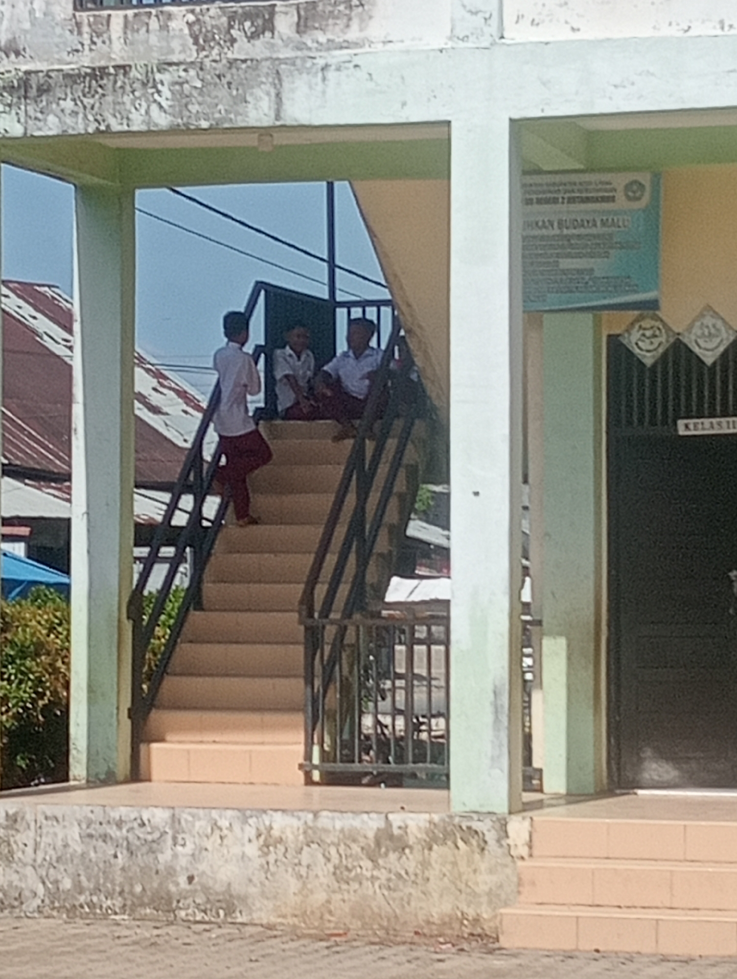 This image captures an outdoor scene at what appears to be a school or educational building in Keudekrueng, Indonesia, under bright daylight. Three young males, dressed in white shirts and dark trousers (one in reddish-brown pants), are present on a concrete staircase with black metal railings. Two boys are seated on the upper steps, seemingly resting or socializing, while another boy stands on a lower step, leaning against the railing.
The building structure consists of light-colored columns and beams. To the right, a dark-colored door is visible, with a white sign above it clearly indicating "KELAS II" (Class II), reinforcing the school setting. Adjacent to the door, on a light yellow wall, hangs a blue-green rectangular sign. Partially legible text on this sign includes "KABUPATEN KOTA" at the top and "EH KAN BUDAYA MALI" below, possibly indicating a local government or cultural program.
In the background, beyond the building's open structure, corrugated metal roofs of other buildings are visible against a clear sky, suggesting sunny weather. The concrete surfaces of the building and stairs show some weathering and discoloration. The overall impression is of a casual moment during school hours in a tropical climate.