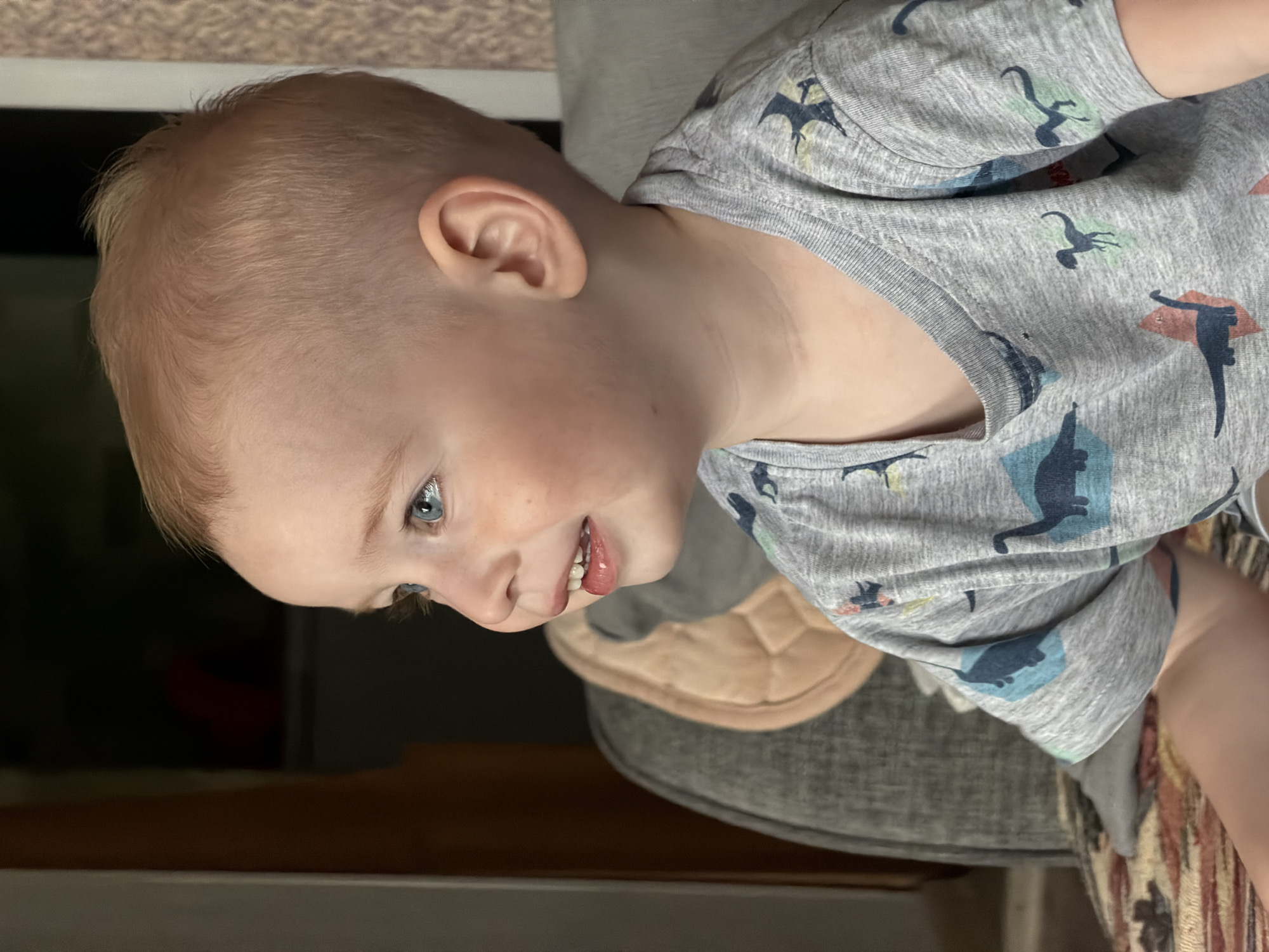A close-up, sideways shot focuses on a young child with light blond hair and blue eyes. The child is wearing a gray t-shirt adorned with a pattern of blue and green dinosaurs. Their head is tilted, with their face looking to the left of the frame. The child's mouth is slightly open, revealing teeth and a hint of their tongue. The background is blurred, showing indistinct shapes and colors that suggest an indoor setting. A quilted tan object rests behind the child's neck, and a portion of a gray chair is visible. The lighting appears to be artificial, casting a warm glow on the child's skin. The overall mood is casual and intimate. No text is visible in the image.