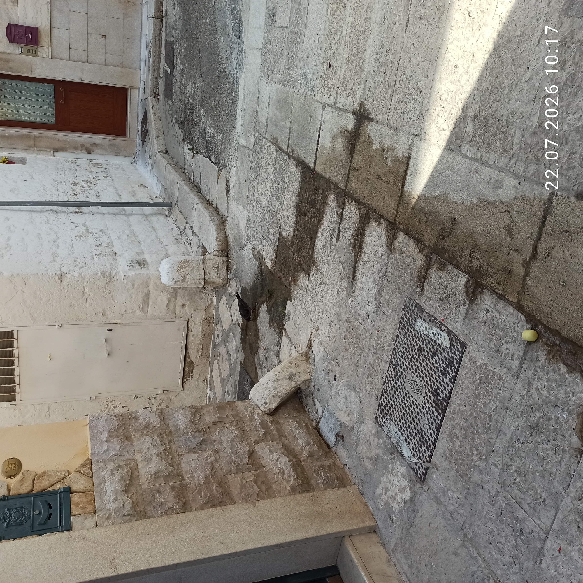 The image depicts a narrow cobblestone street in Modugno, Italy, on a sunny day, as indicated by the timestamp "22.07.2026 10:17". The street is lined with old stone buildings. On the left, a light-colored stone wall with a dark green mailbox is visible. Further down the street, a white metal door is set into a building. On the right, a wooden door with a dark frame is present, and a purple mailbox is mounted on the wall beside it. A bird is seen drinking from a puddle on the street. The ground is paved with large, irregular stone slabs, some of which are wet and discolored, suggesting recent water spillage or drainage. A metal grate is visible on the ground in the foreground, and a small yellow object, possibly a piece of fruit, lies on the paving stones. The overall scene conveys a sense of everyday life in an older European town.