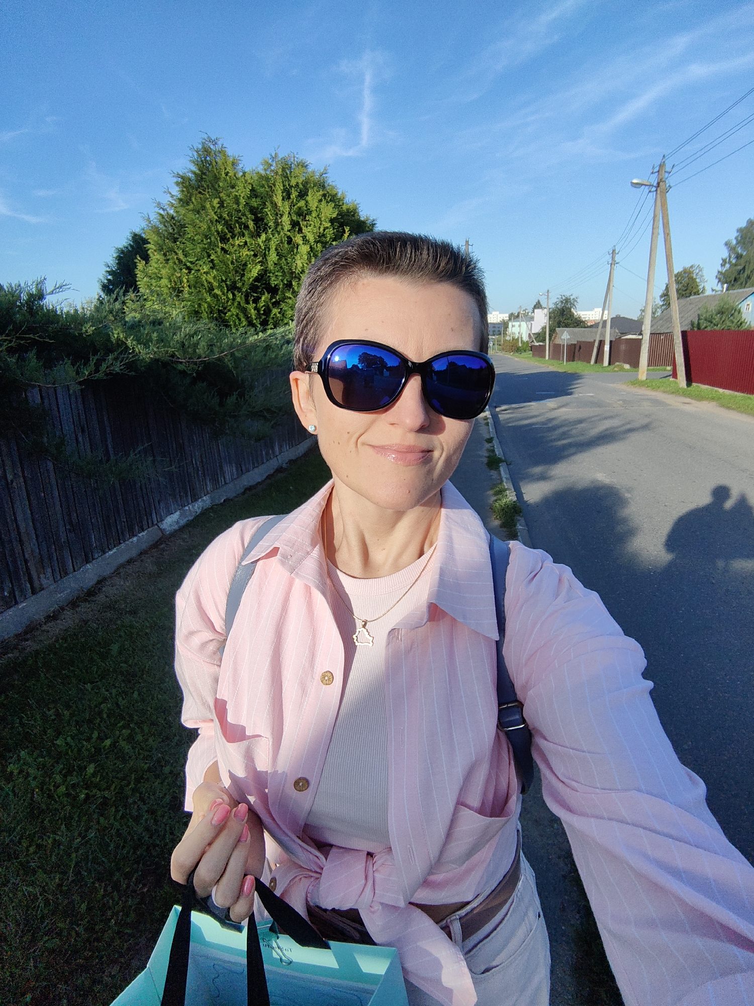 A woman with short, dark hair and sunglasses is taking a selfie outdoors. She is wearing a light pink striped button-down shirt over a light pink ribbed top, light colored pants, and a necklace with a pendant. She is holding a turquoise shopping bag in her left hand. The woman is standing on a paved road with a grassy shoulder and a wooden fence with trees behind it on her left. In the background, there are houses with red fences, utility poles, and a clear blue sky. The scene appears to be in a suburban area of Fanipal, Belarus.