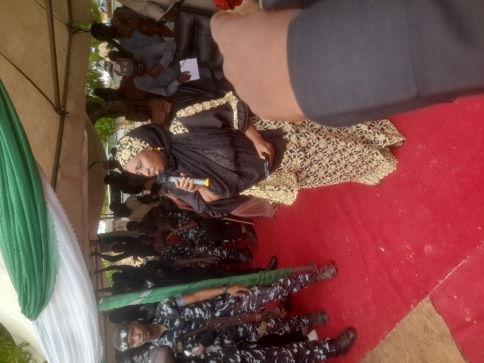 A woman in a black and gold dress speaks into a microphone. She is under a tent with green and white decorations. Behind her, a crowd of people sits in rows. In the foreground, uniformed officers lie on the ground with rifles. The ground is red. The image is taken at an angle.