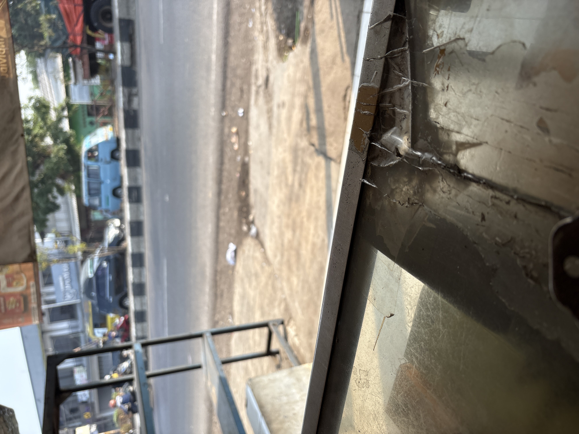A slightly out-of-focus, eye-level shot captures a street scene in Palimanan, Indonesia, viewed from behind a covered stall. In the foreground, a scratched and worn metallic surface, likely a counter or table, dominates the lower portion of the frame. The background reveals a busy street with traffic including a blue van, several motorcycles, a black SUV, and a red truck. Buildings line the street, some with awnings, and trees are visible behind them. The road surface is paved, with a striped pedestrian crossing. The overall lighting suggests daytime, possibly late afternoon due to the warm tones and shadows.