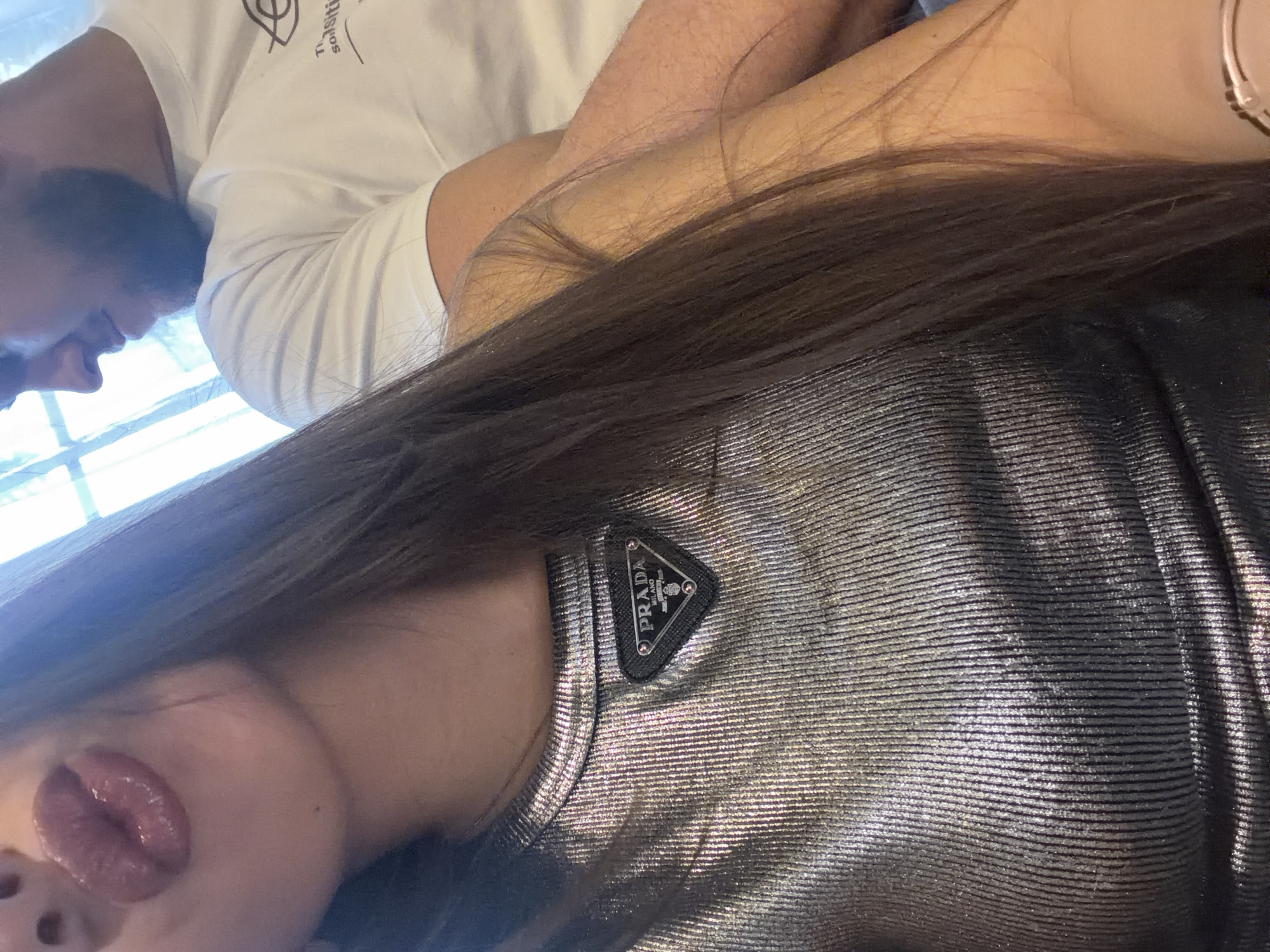 A close-up, tilted shot shows a woman with long, dark hair wearing a silver top with a Prada logo. Adjacent to her, a man with a beard wears a white t-shirt. The setting appears to be indoors, possibly near a window given the natural light. The location is Rudartsi, Bulgaria.