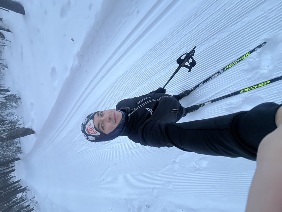 An adult male is performing a selfie while cross-country skiing on a groomed snow trail in a forested area of Kazan, Russia. The subject, positioned centrally, looks towards the camera with a neutral expression. He wears a dark winter jacket with a zipper and white accents, a dark neck gaiter, and a dark winter hat featuring white markings and Cyrillic text, "АК БАРС". His right arm (viewer's left) is extended, holding the camera. His left hand holds a black ski pole. He stands on two black cross-country skis clearly branded "FISCHER". The ski track ahead and behind him displays distinct parallel grooves for classic skiing, flanked by a wider, flatter section. The environment is a dense, snow-covered forest with numerous trees under an overcast sky, indicating winter conditions.